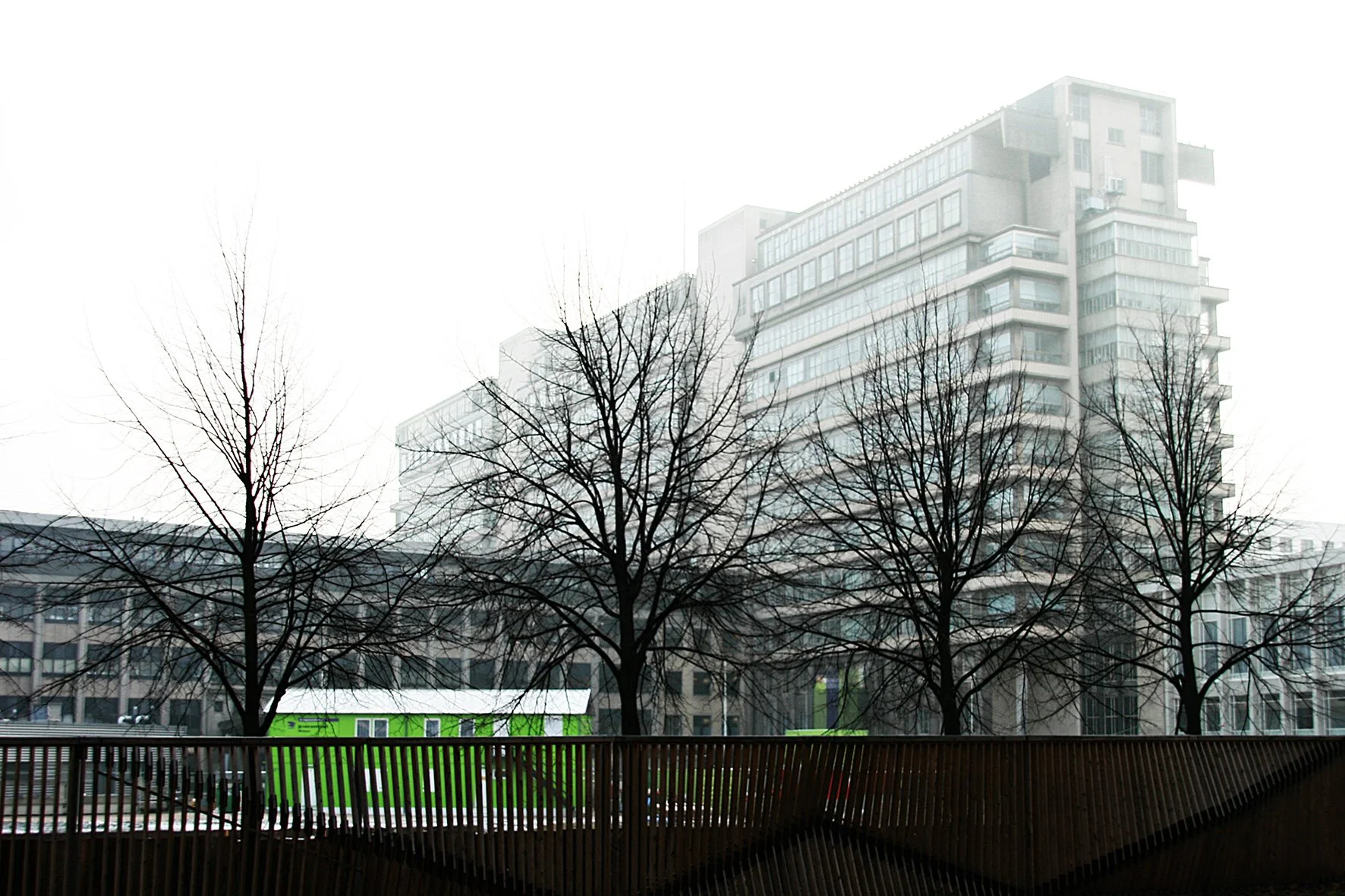 Vue d'une ville automnale avec des arbres sans feuilles, un bâtiment moderne en arrière-plan, et une structure en bois au premier plan, sous un ciel gris.