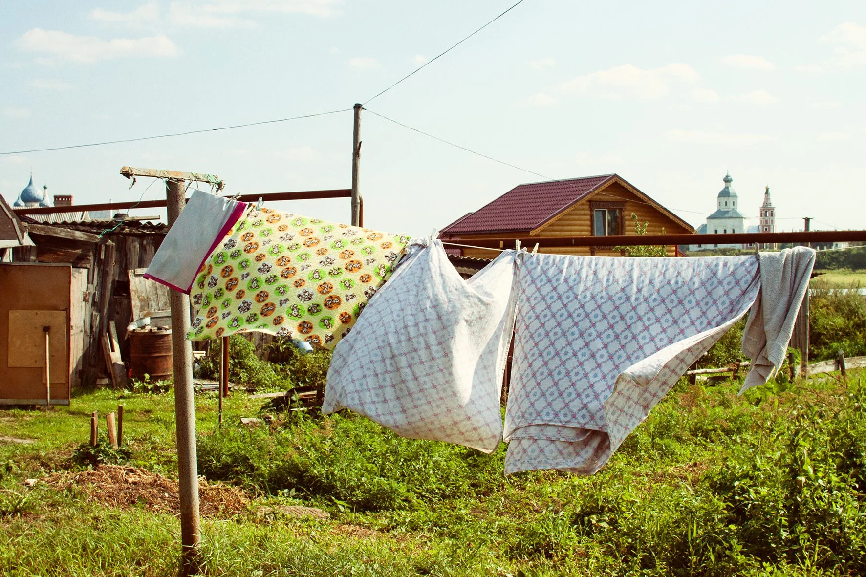 Linges à sécher suspendus sur une corde dans un jardin rural avec des bâtiments en bois, et des églises en arrière-plan.