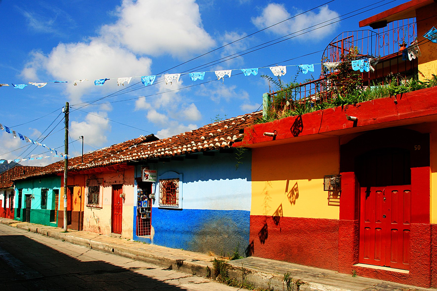Maisons colorées avec façades en rouge, bleu, vert et jaune dans un village, sous un ciel bleu avec des nuages, décorées de banderoles bleues et blanches.