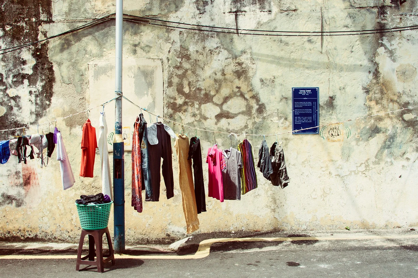 Vêtements suspendus à une corde en face d’un mur usé, avec une poubelle remplie de vêtements et un panneau bleu sur le mur.