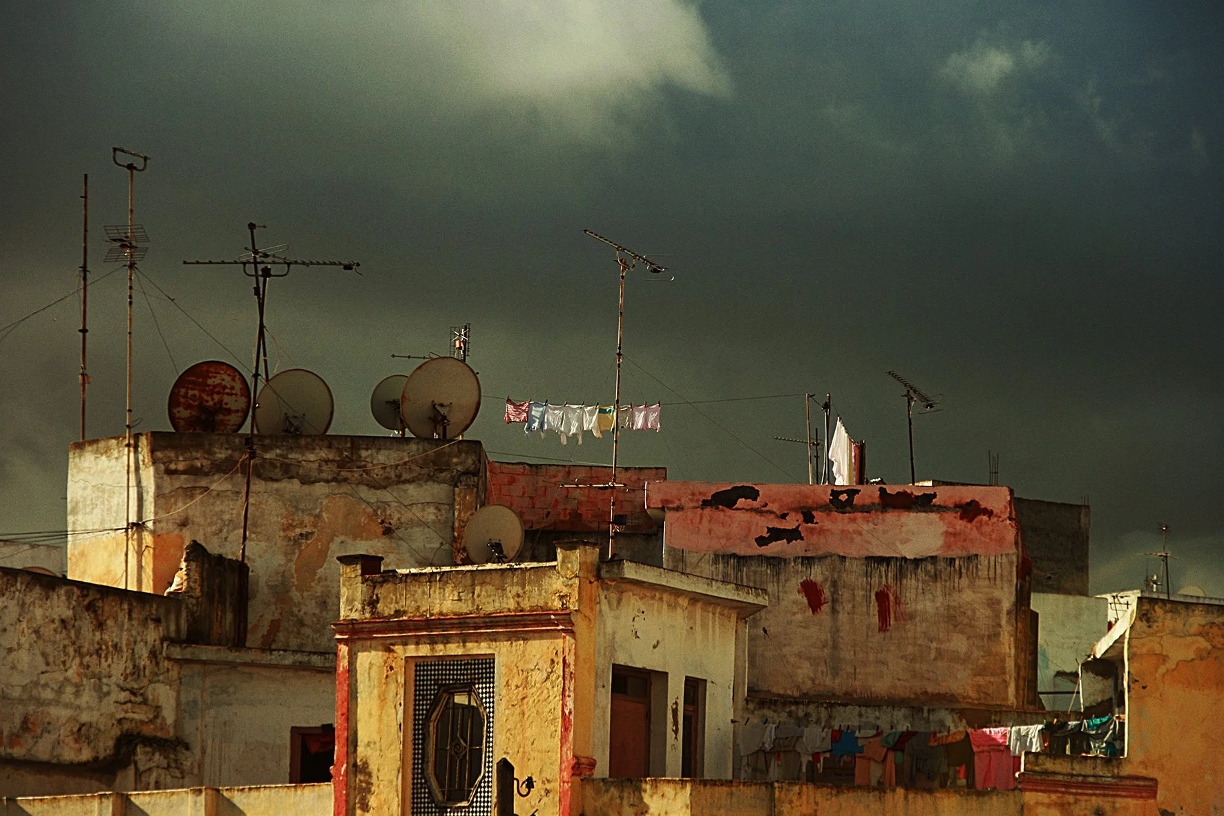 Toits d'immeubles délavés avec des antennes, des paraboles et du linge suspendu, sous un ciel nuageux.