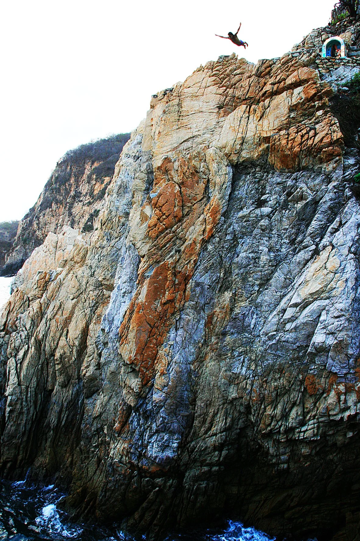 Une personne saute en haut d'une falaise rocheuse au bord de l'océan, avec une maison au sommet.