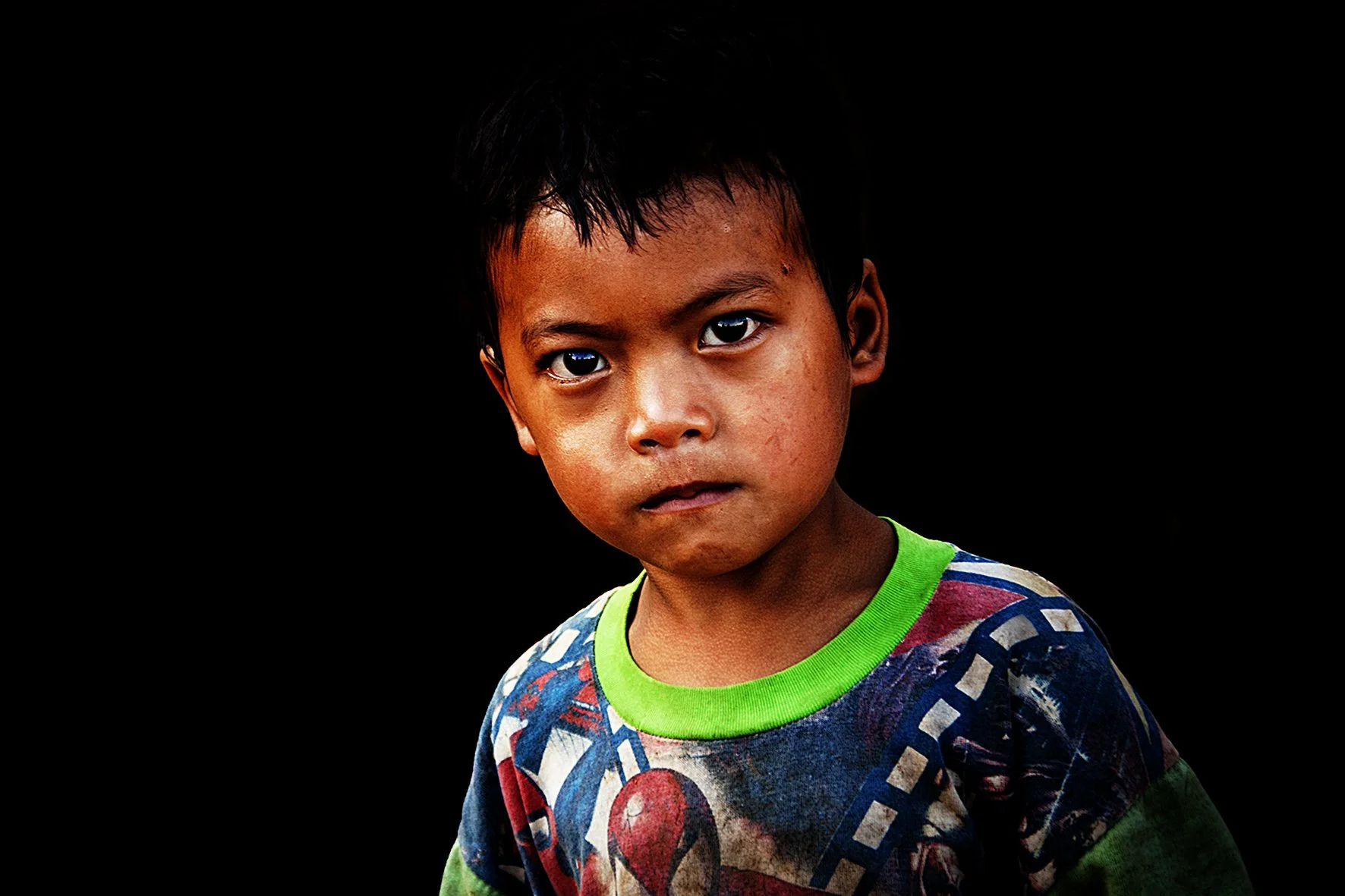 Portrait d'un jeune garçon aux yeux sombres, portant un t-shirt coloré, contre un fond noir.