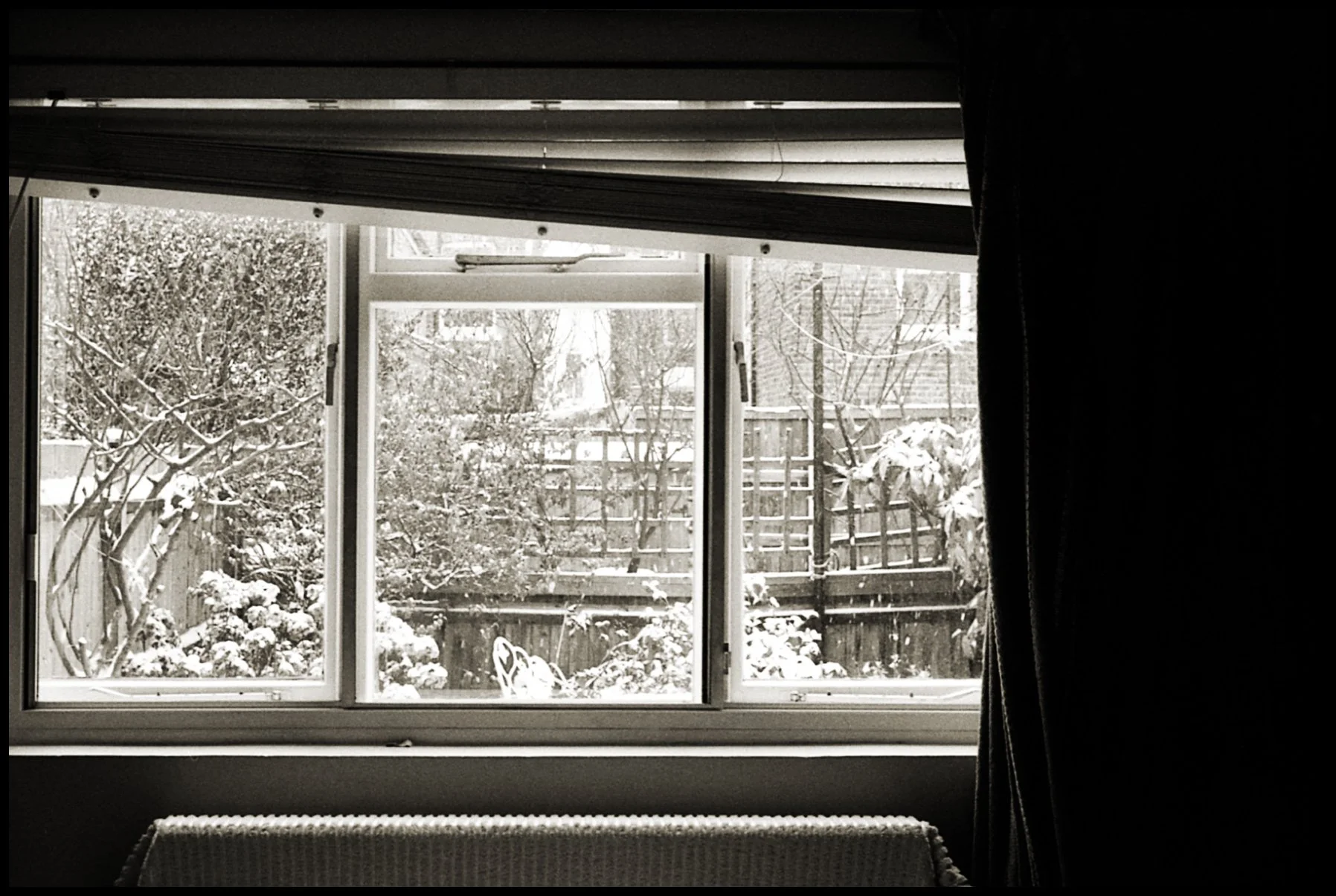 Vue d'une fenêtre ouverte donnant sur un jardin enneigé avec des arbres et une clôture en bois, vue de l'intérieur d'une pièce sombre avec une chaise ou un banc en bois.