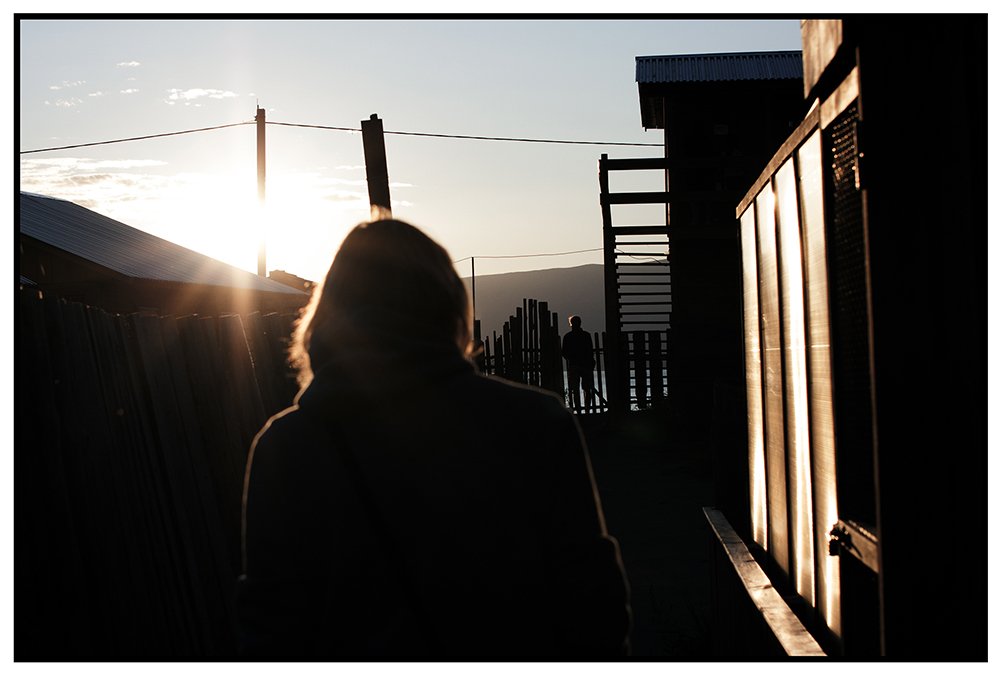 Silhouette d'une personne dans un couloir extérieur, avec la lumière du soleil à l'arrière-plan, montrant un paysage de montagnes au loin, avec d'autres personnes visibles en arrière-plan.