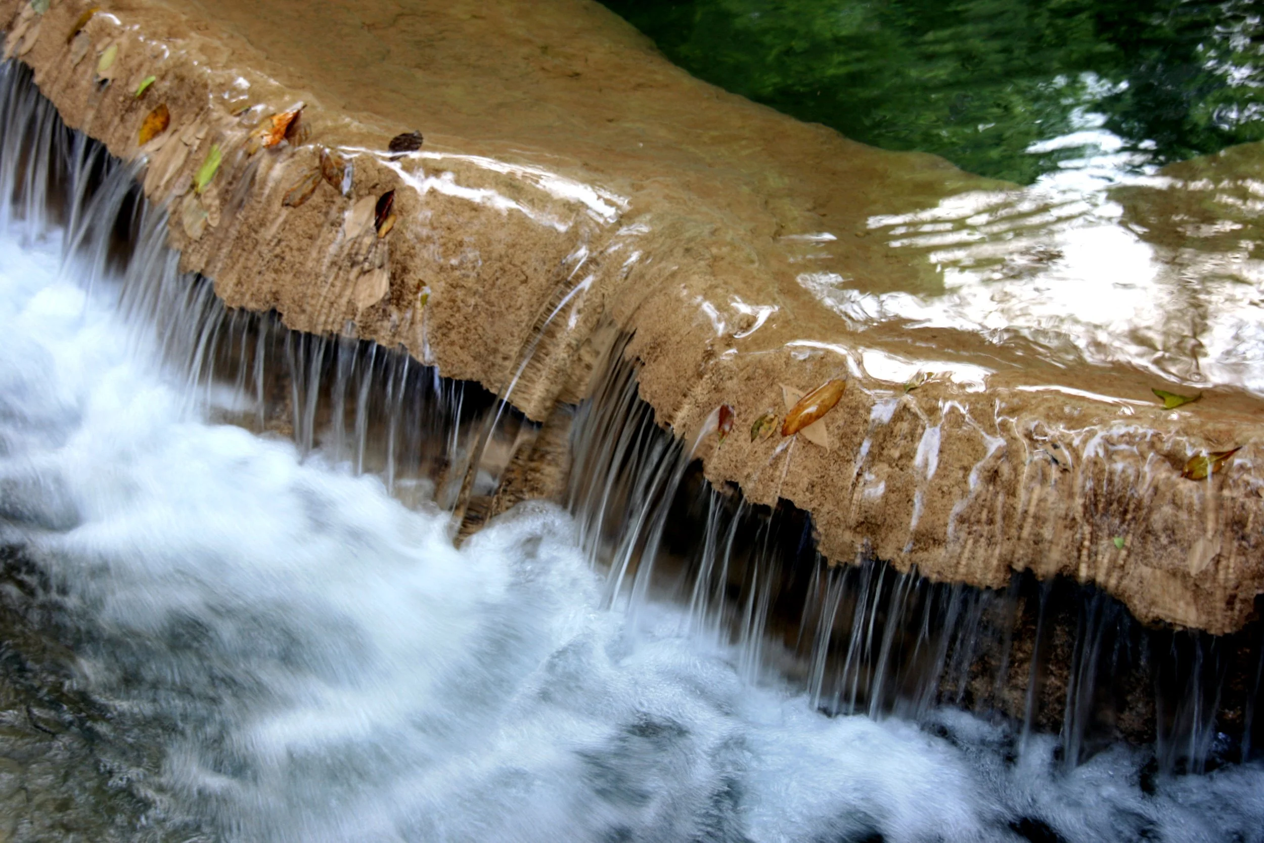 Petite cascade d'eau tombant sur une roche, avec des feuilles dispersées, dans un environnement naturel.