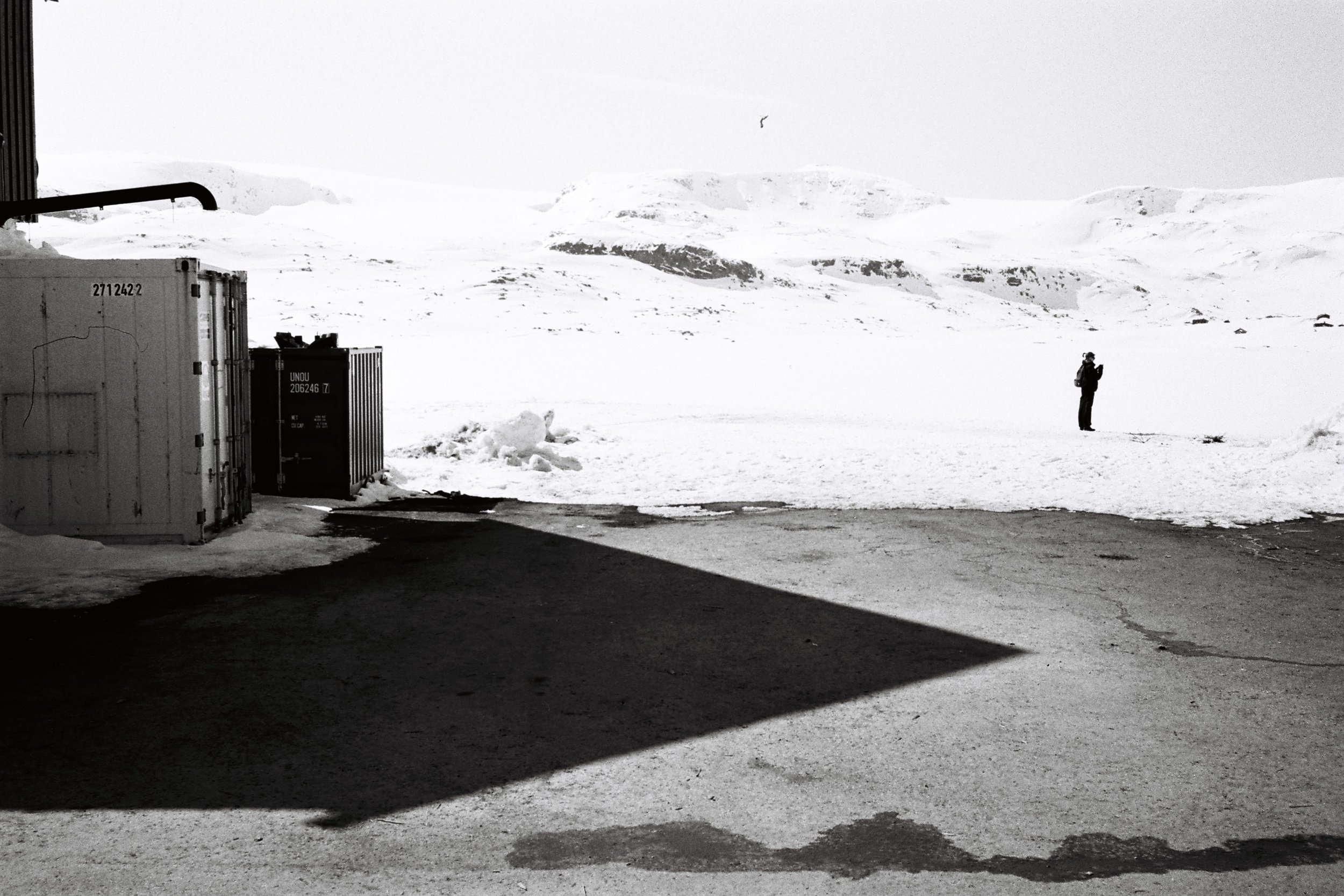 Vue d'une zone enneigée avec un homme debout au loin, containers de stockage et ombres au premier plan, paysage montagneux en arrière-plan