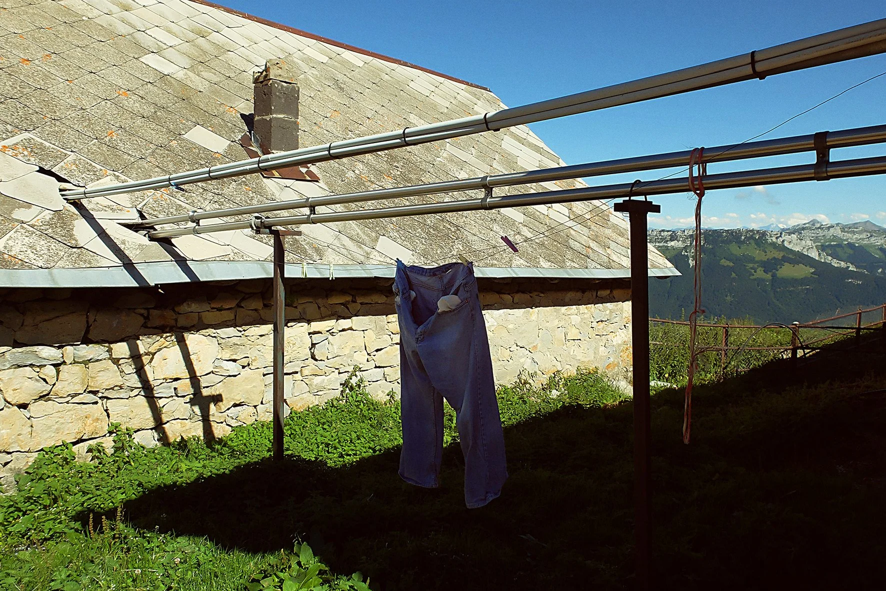 Une paire de jeans suspendue à un fil à linge, avec une maison en pierre et un paysage montagneux en arrière-plan.