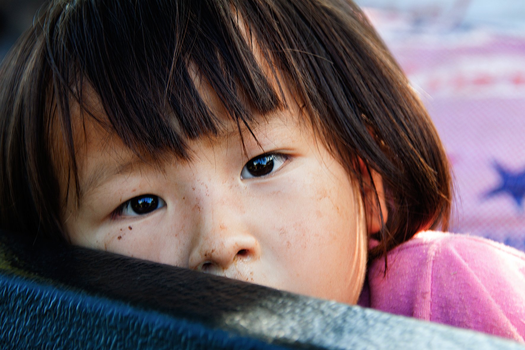 Jeune fille aux cheveux bruns, yeux marron, regardant vers l'objectif, portant un haut rose, face partiellement cachée derrière une surface noire.