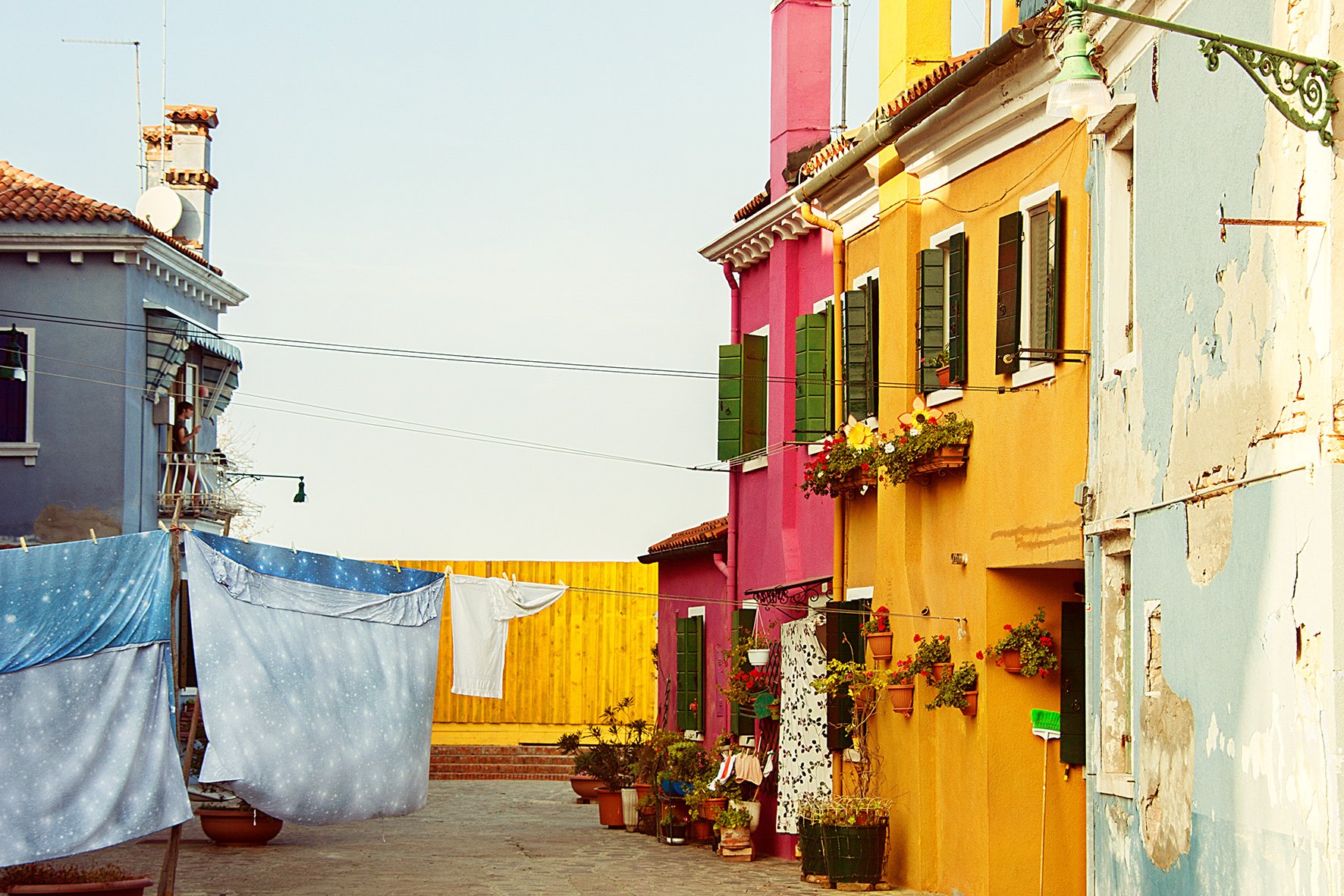 Maisons colorées avec volets verts, fleurs sur les fenêtres, linge suspendu à des cordes, dans un quartier ensoleillé.