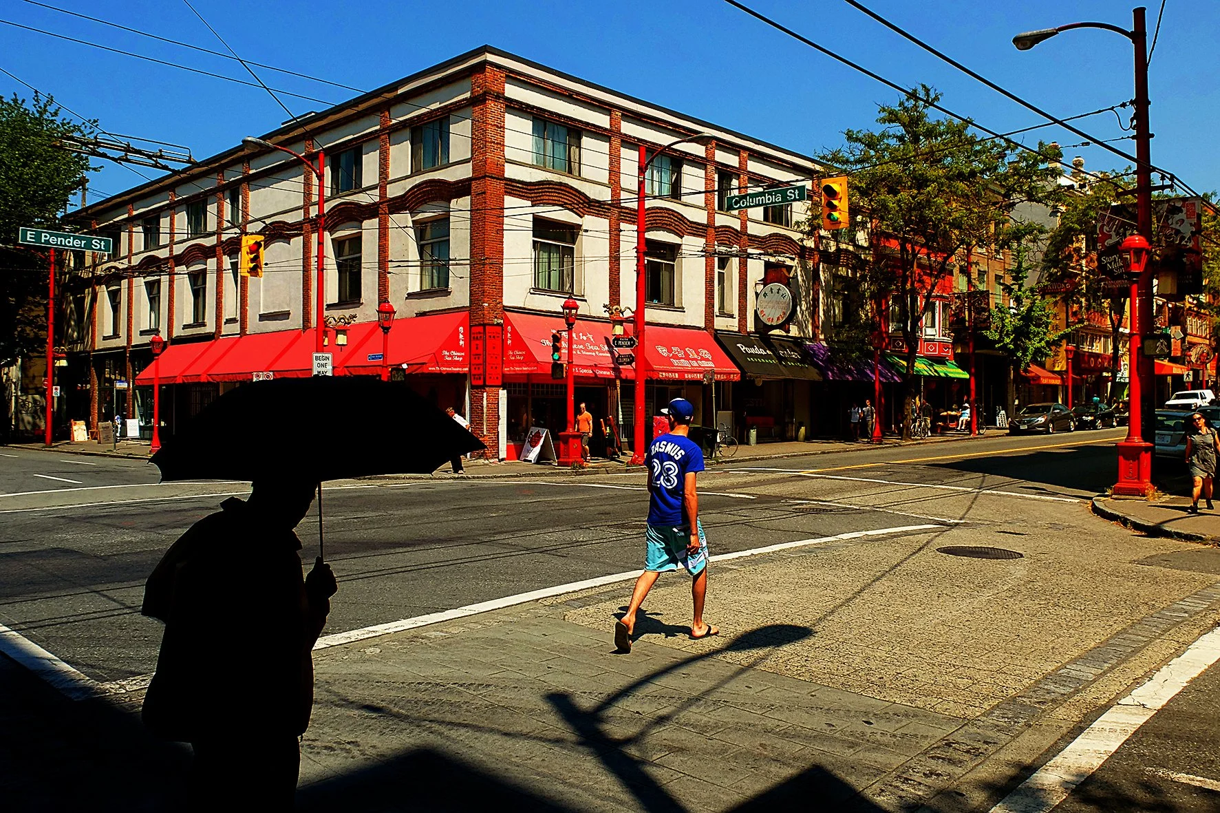 Une rue animée avec un bâtiment à étages abritant des boutiques, une personne marche avec un parapluie en silhouette dans l'ombre, une autre personne traverse la rue en portant un maillot de sport. La scène est en plein jour avec un ciel bleu clair.