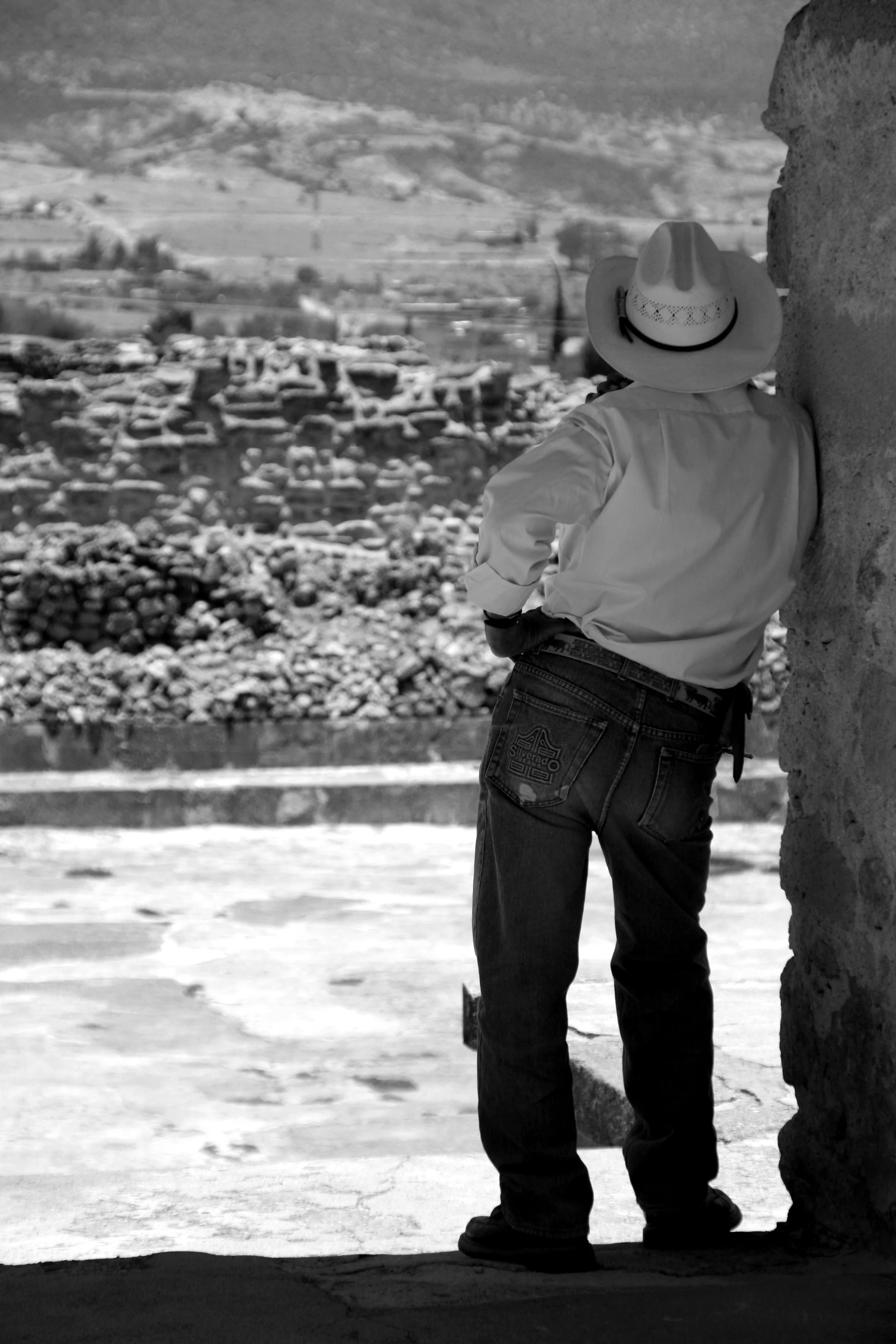 Un homme portant un chapeau de cowboy regarde au loin à travers une ouverture dans un bâtiment en pierre, avec un paysage rural en arrière-plan.