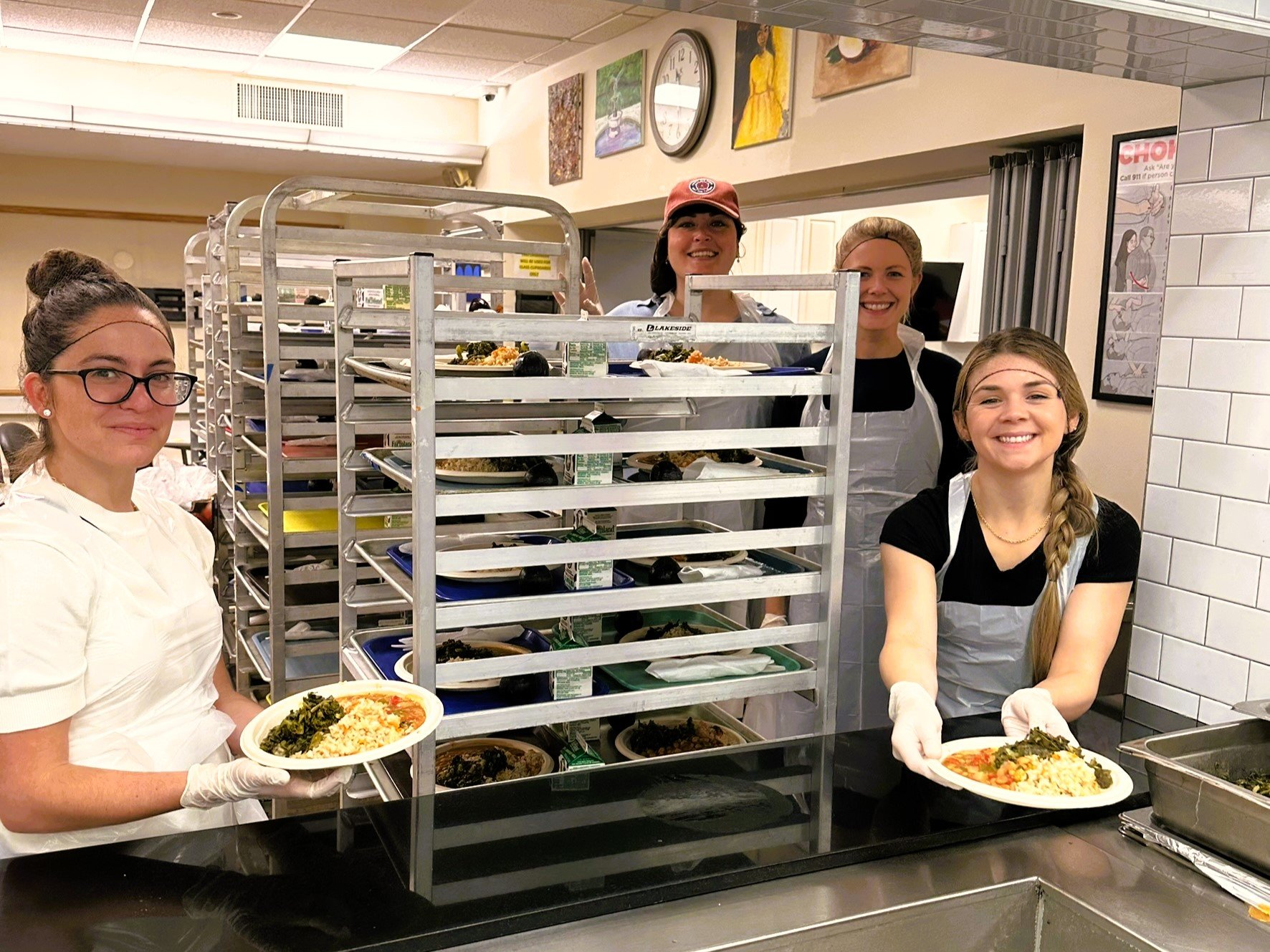 photo of four young women holding plates of freshly cooked meals in a kitchen