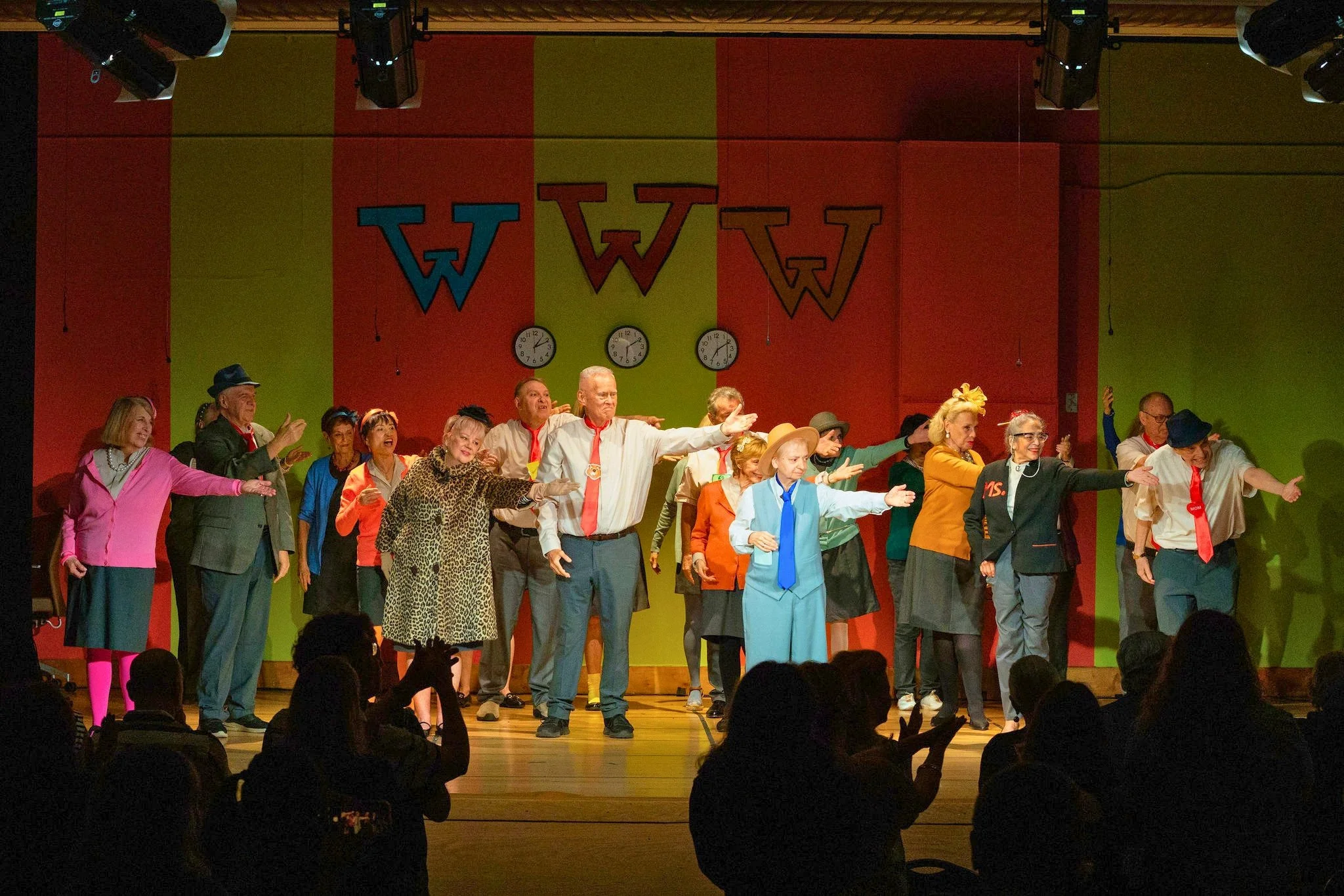 older adults on an auditorium stage in business attire costumes waving their left arms to an unseen person offstage