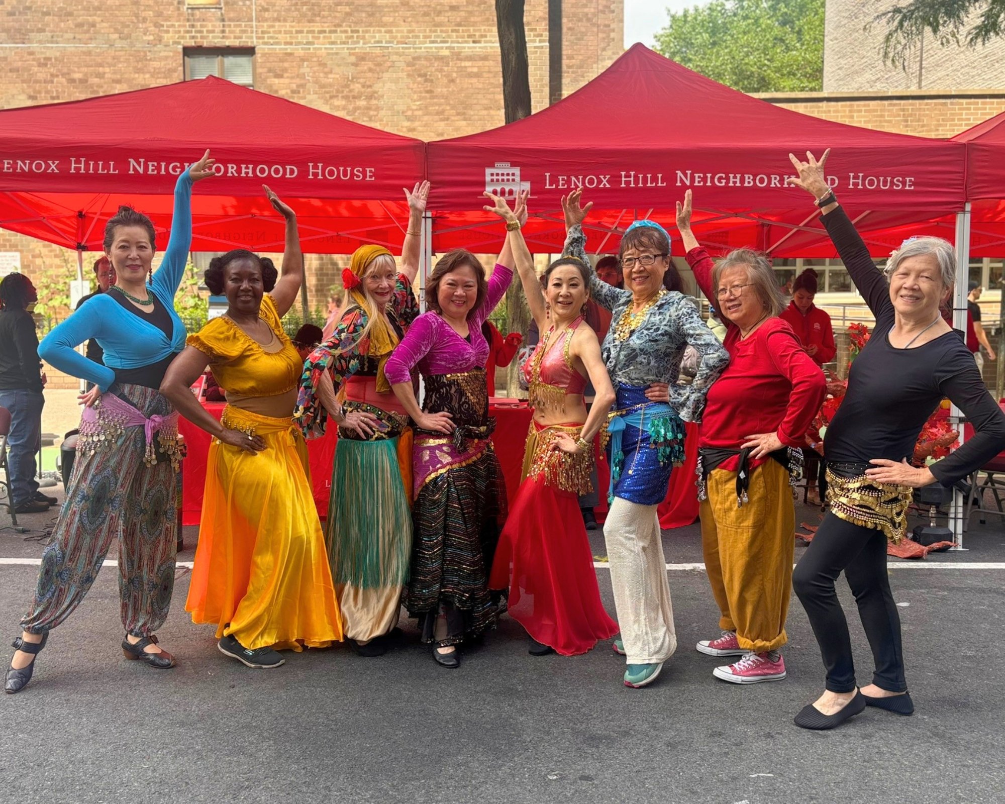 image of 8 women in belly dancing outfits posing in front of a red tent that reads "Lenox Hill Neighborhood House" in white font