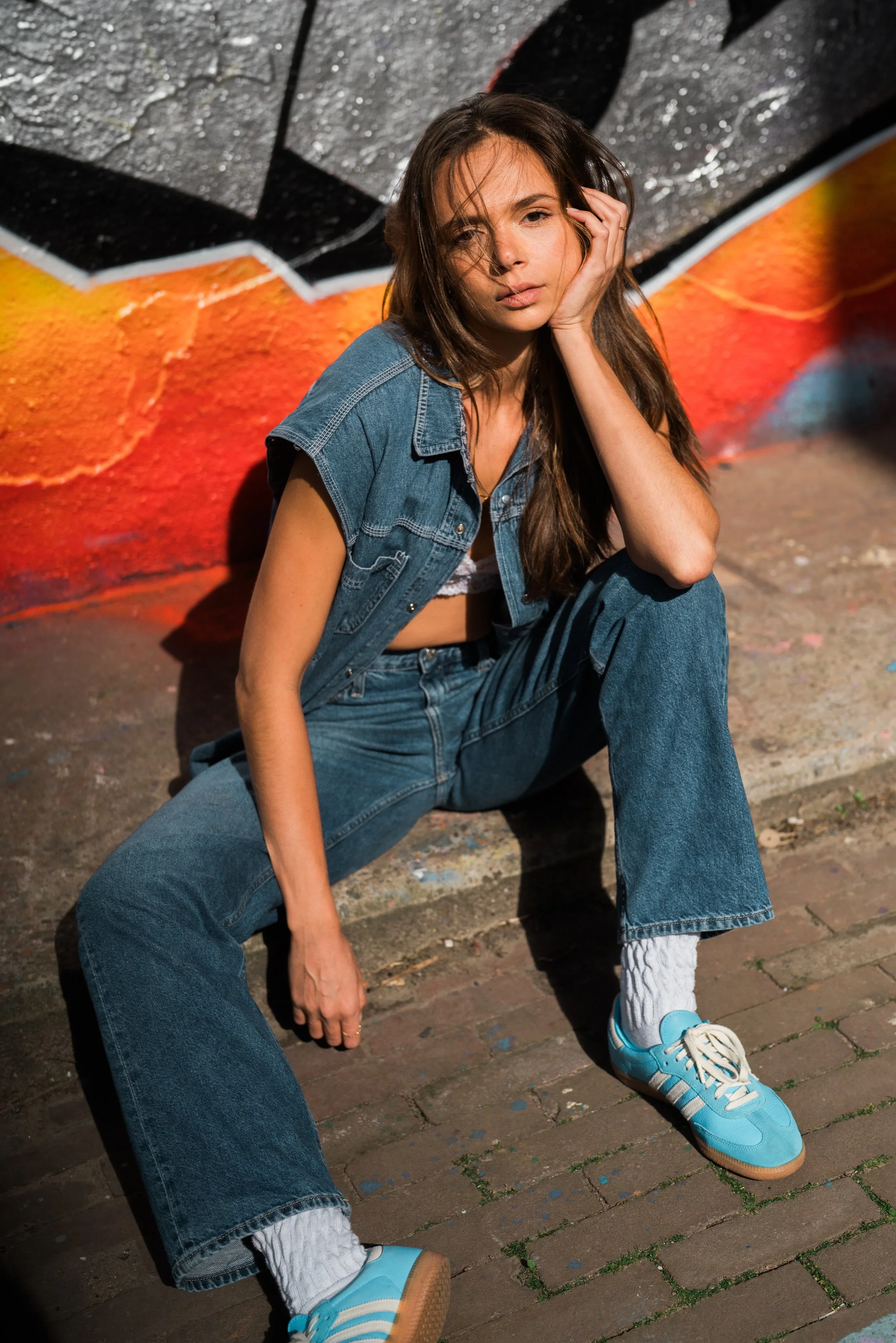 A young woman with long brown hair, wearing a sleeveless denim jacket, high-waisted jeans, white socks, and blue sneakers, sits on a brick sidewalk resting her head on her hand against a colorful graffiti wall.