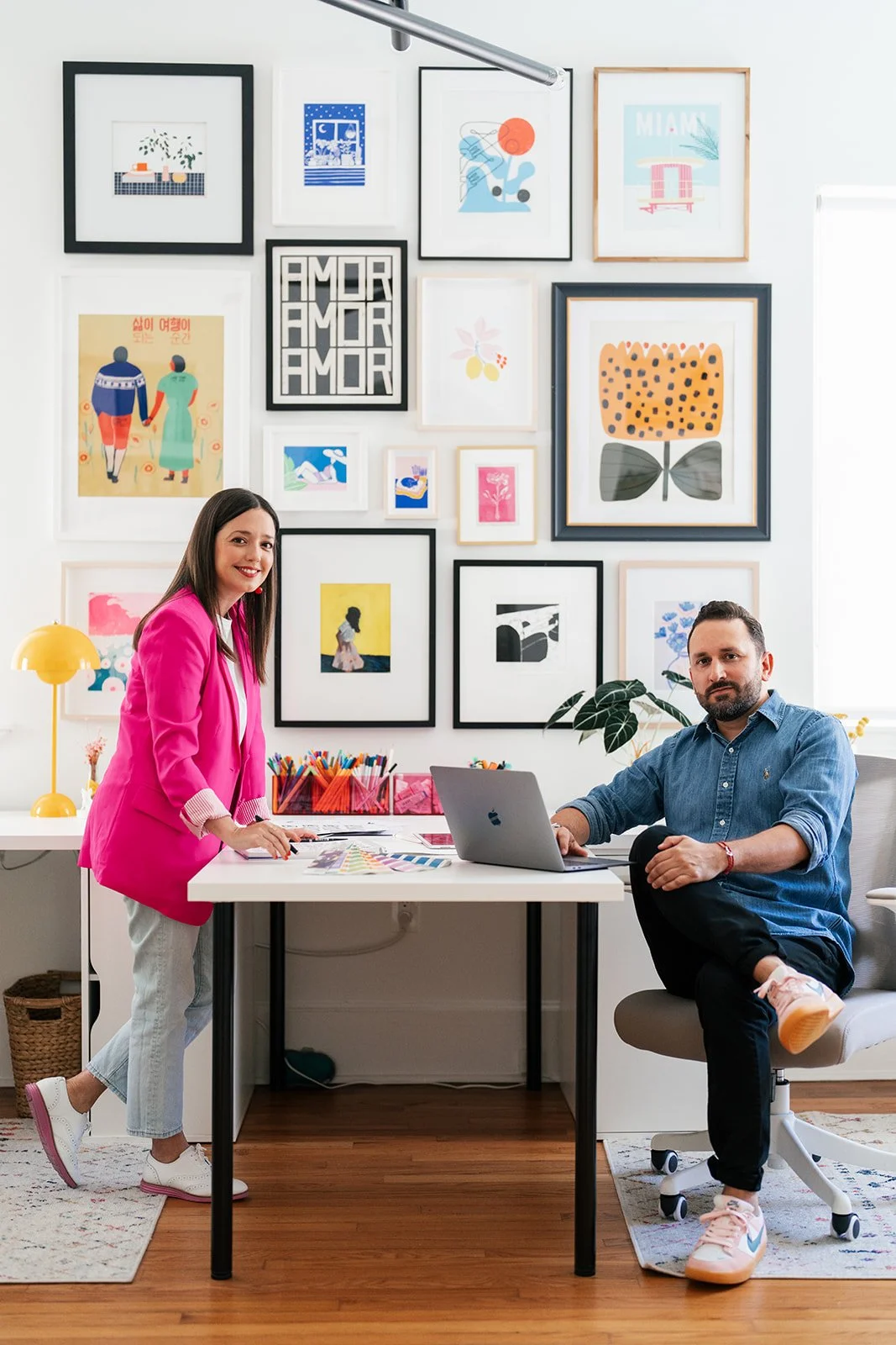 A woman in a pink blazer and white sneakers standing at a white table, and a man in a blue shirt and black pants sitting in a gray office chair in a creative workspace with colorful framed artwork on the wall.