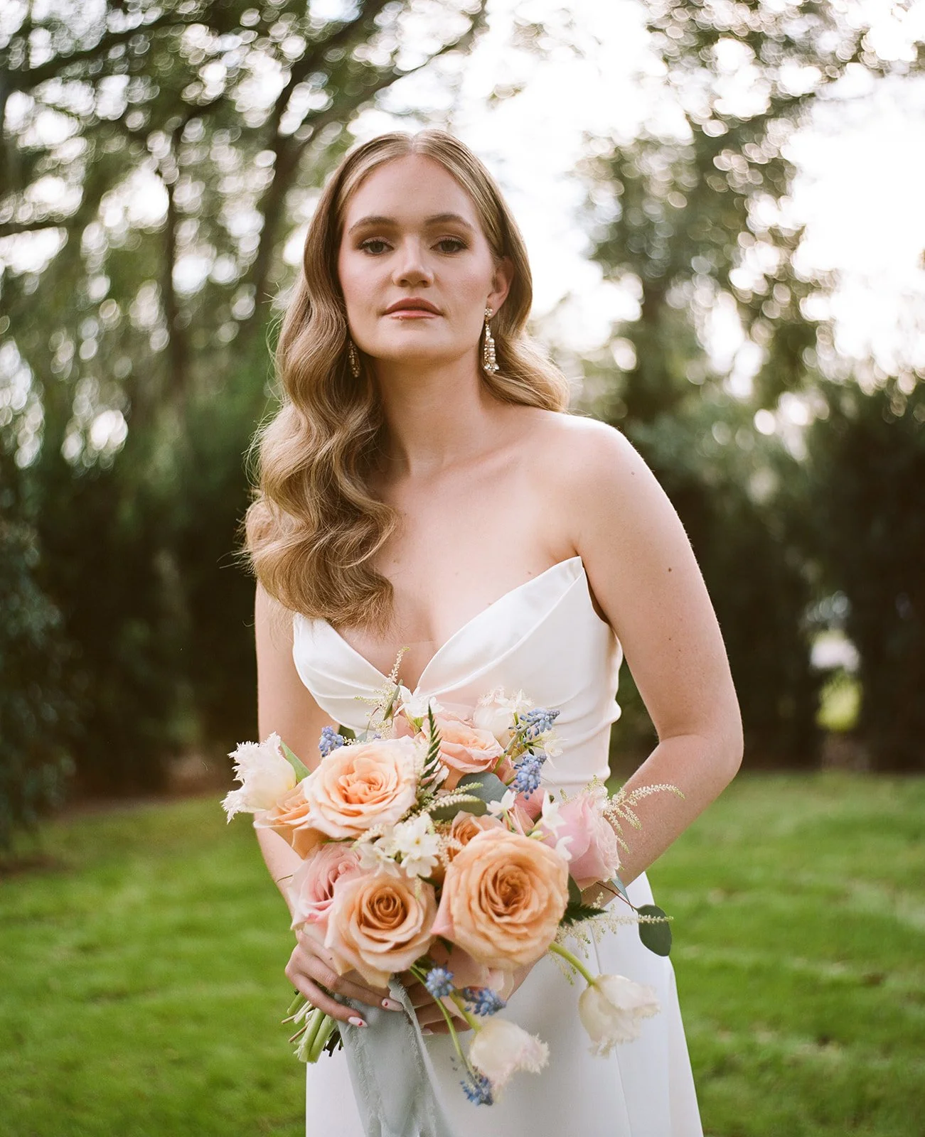 A bride holding a bouquet of pink and white roses with blue accents, standing outdoors with trees in the background.