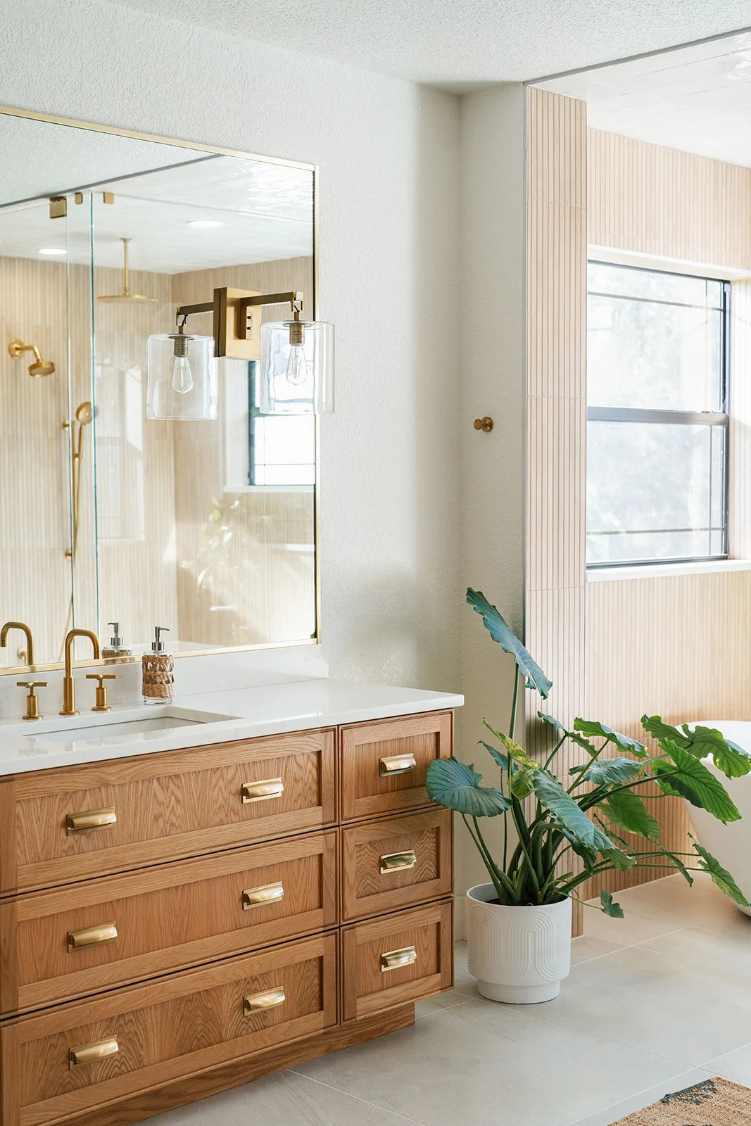 A bathroom vanity with a large mirror, gold fixtures, and a wooden cabinet. There is a large potted plant next to the vanity, near a window with natural light.
