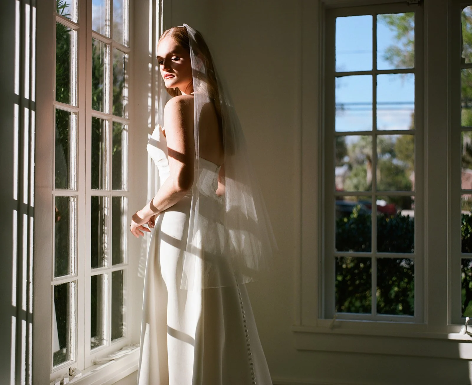 A bride standing by a window with sunlight casting shadows on her, wearing a wedding dress and veil.