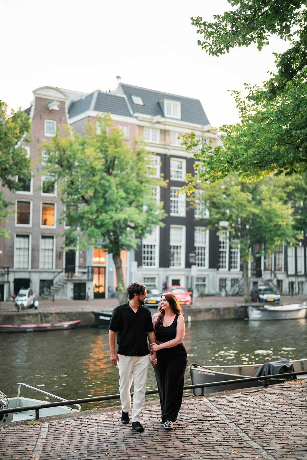 A couple walks hand in hand along an Amsterdam canal with houses and trees in the background during sunset.