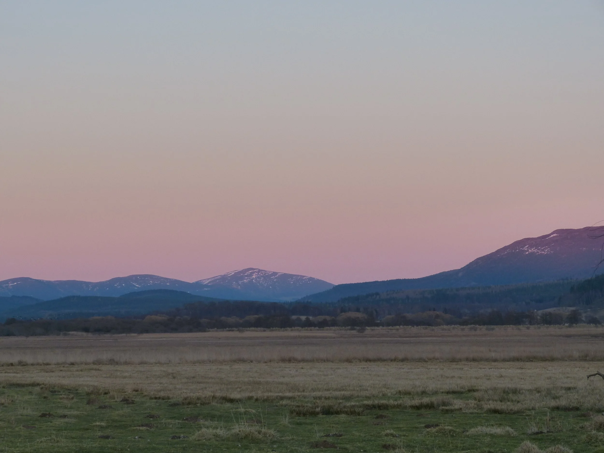 Scenic landscape of distant mountains with snow on the peaks, under a sky with a gradient from pinkish near the horizon to blue higher up, taken during twilight or early evening.