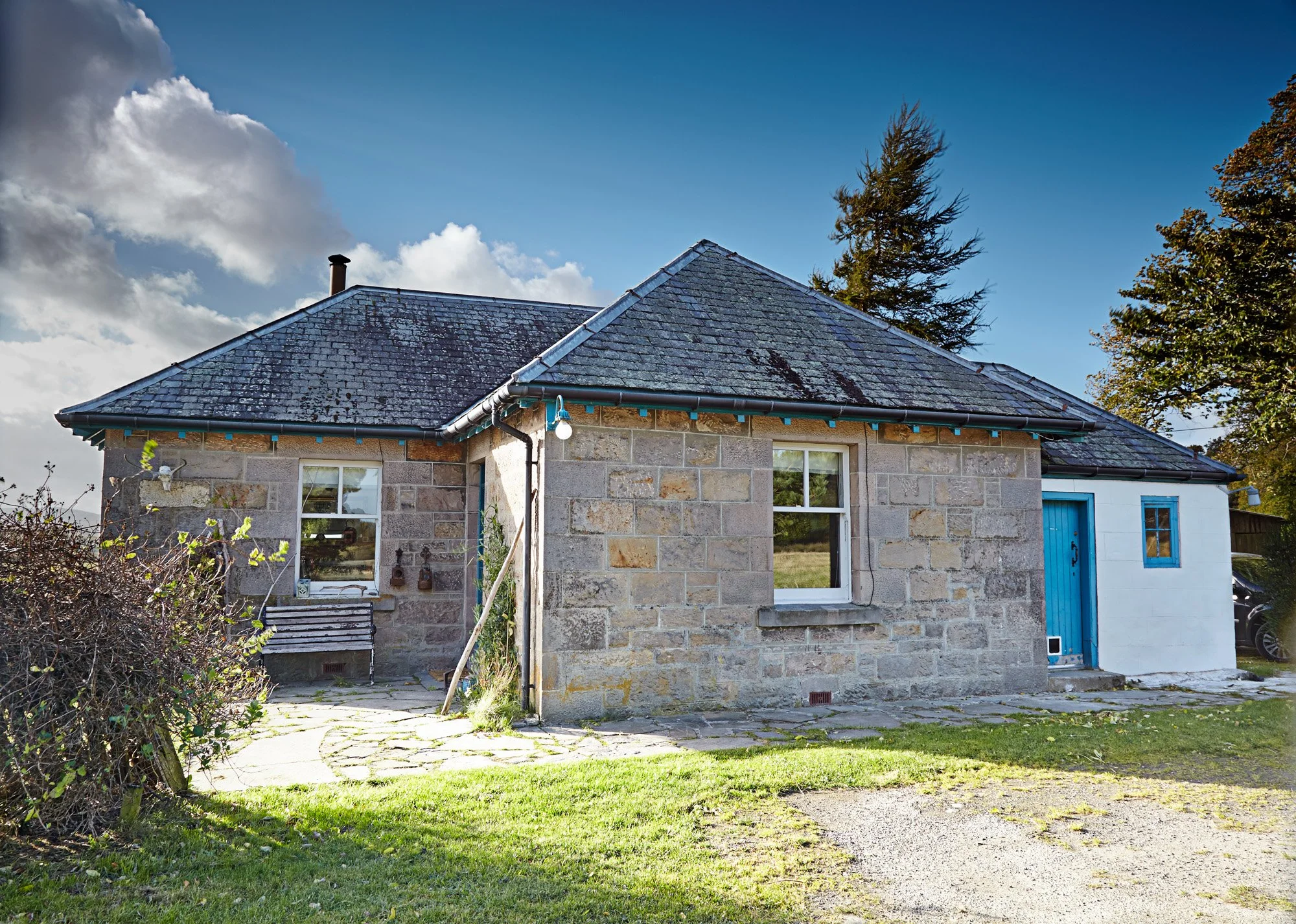 A house with a stone exterior, blue door, and black shingle roof under a partly cloudy sky with trees in the background.