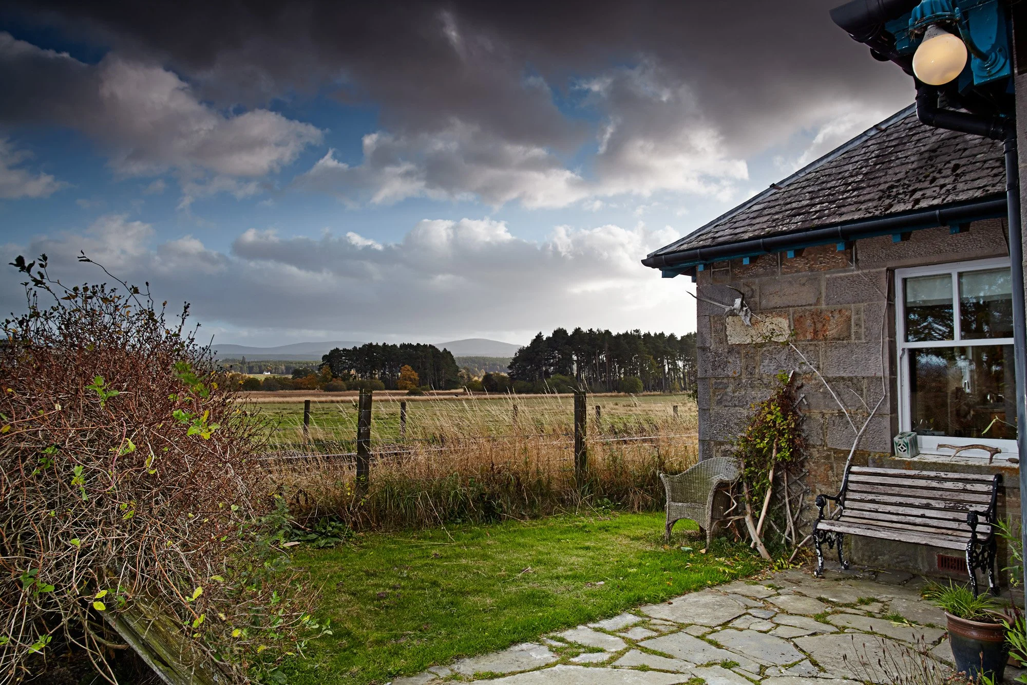 An outdoor scene showing a lawn, a paved patio area, and a stone house with a window and a bench. There is a weathered fence and some bushes in the foreground, and fields and trees in the distance under a partly cloudy sky.