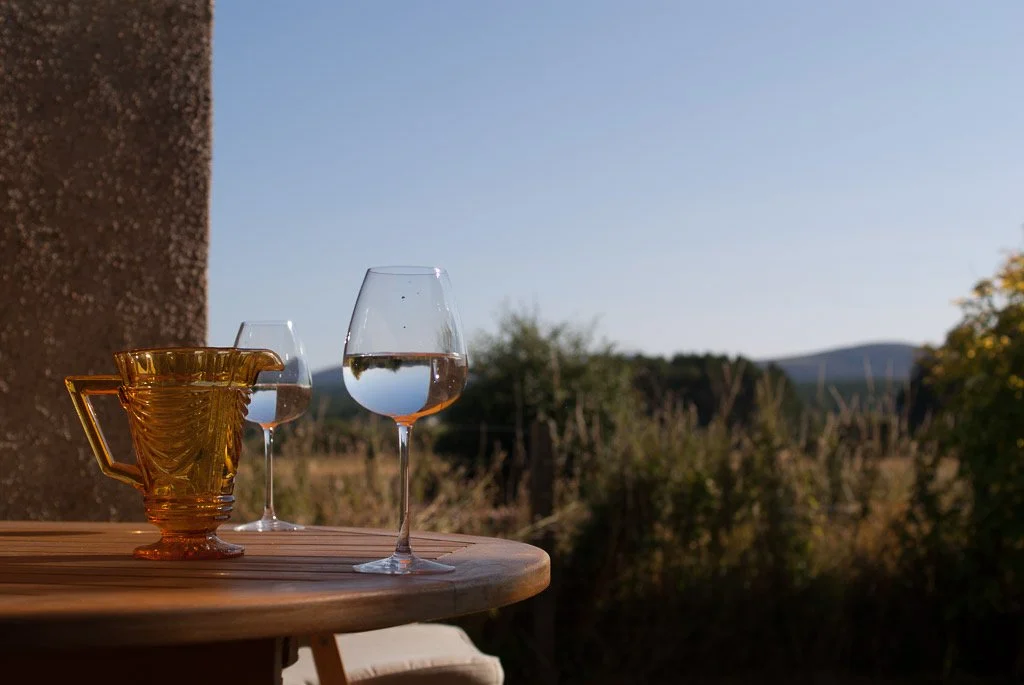 Glass of water, glass of wine, and a yellow glass pitcher on a wooden table outside with a landscape of trees and hills in the background.