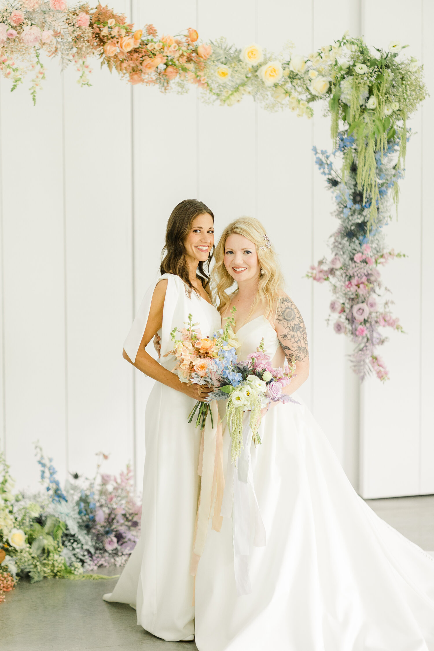 Bride and bride posing for couple portraits holding pastel florals. Bride on the left is wearing the Windham jumpsuit and the bride on the right is wearing Ines Di Santo Quice.