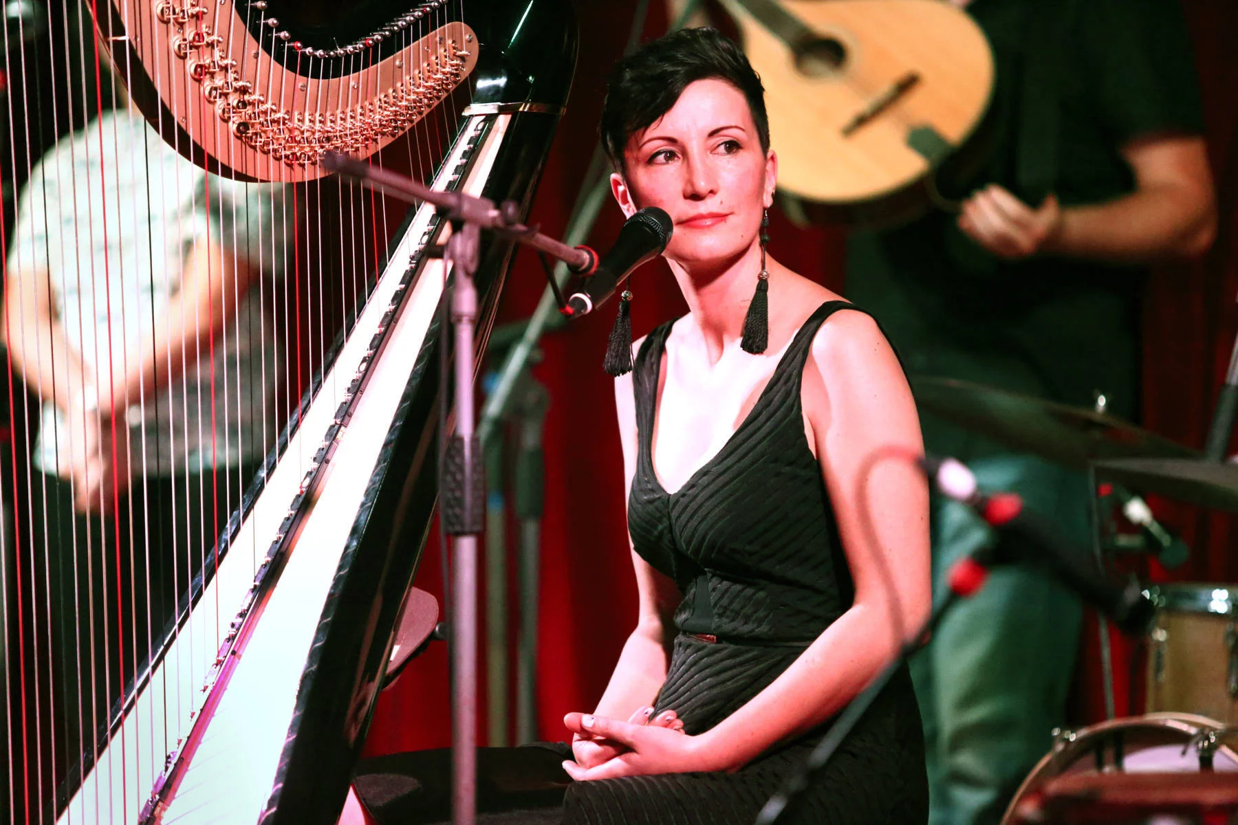 Emily Rosner with short black hair in a black dress sits at a piano with a serious expression. Musicians with guitars and drums are in the background.