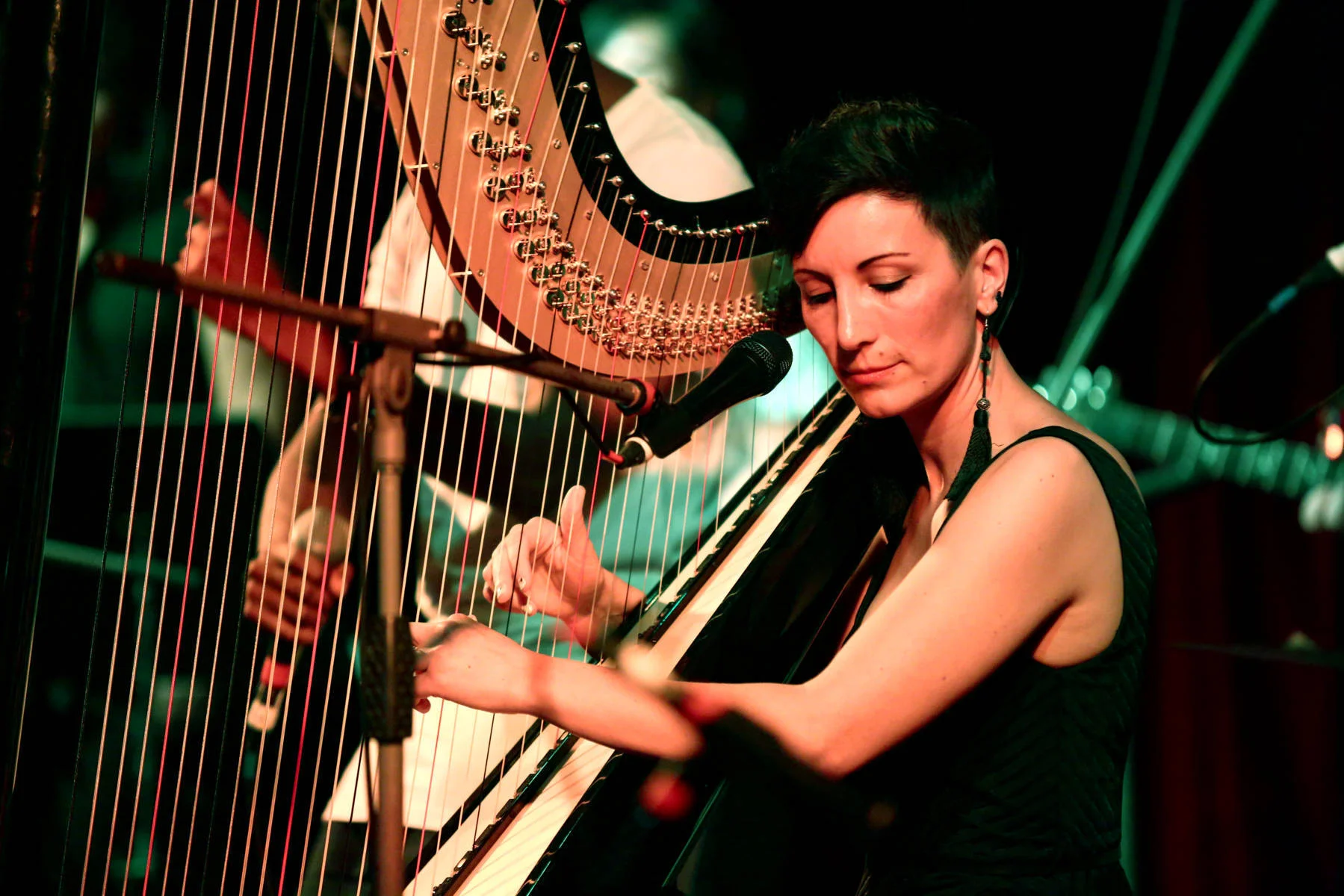 A woman with short dark hair playing a harp and singing into a microphone on stage.