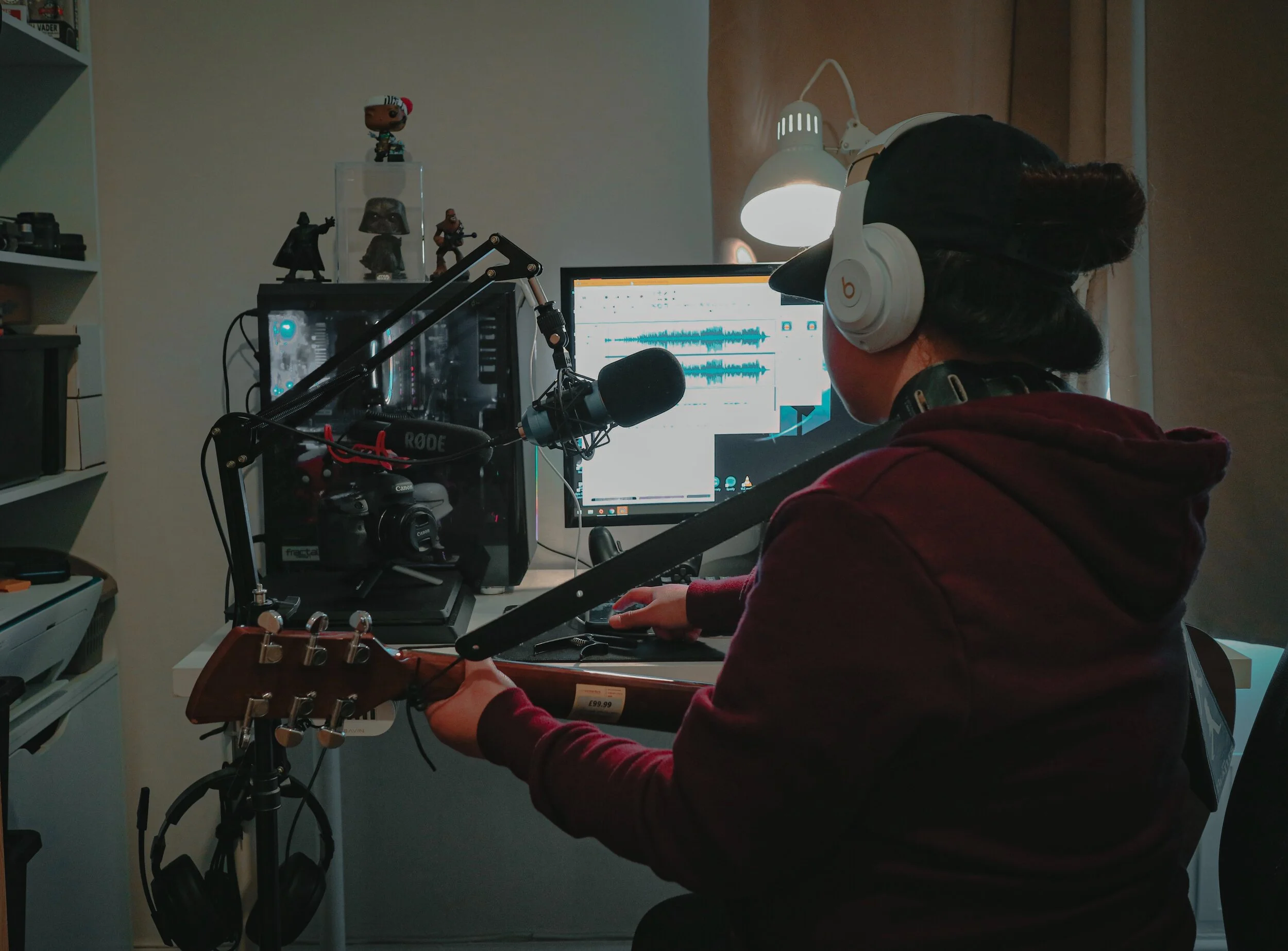A person wearing a maroon hoodie and white headphones, sitting at a desk in front of a computer monitor, recording music or audio with a microphone, in a room with shelves holding figurines and other items.