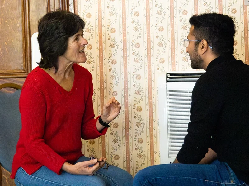 A woman in a red sweater and a man in a black shirt engaging in conversation indoors, seated near a window with patterned wallpaper in the background.