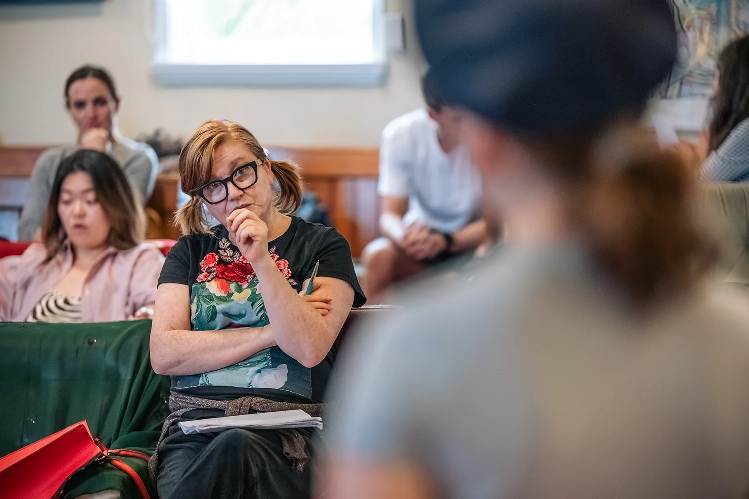 Juliet Furness, an acting coach, sitting and watching students perform in class, listening intently to someone speaking, in a room with other seated people.