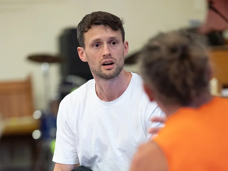 A man with short dark brown hair, wearing a white t-shirt, looks surprised or confused while engaging in conversation with another person in an indoor setting.