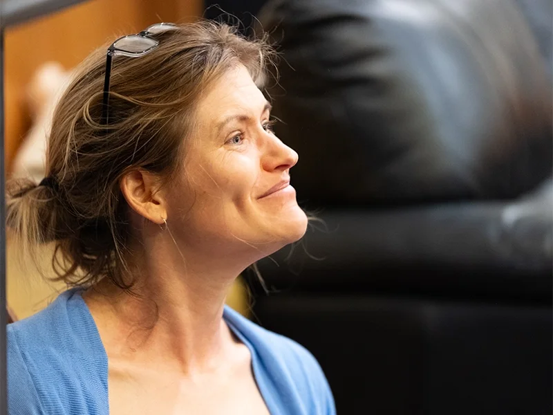 A woman with light brown hair, glasses on her head, smiling and looking to her left, wearing a blue shirt.