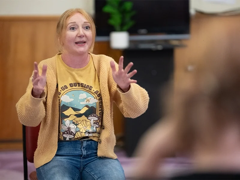 A woman with red hair wearing a yellow t-shirt and a beige cardigan, gesturing with her hands while speaking indoors, with a blurred person in the foreground and a TV and potted plant in the background.