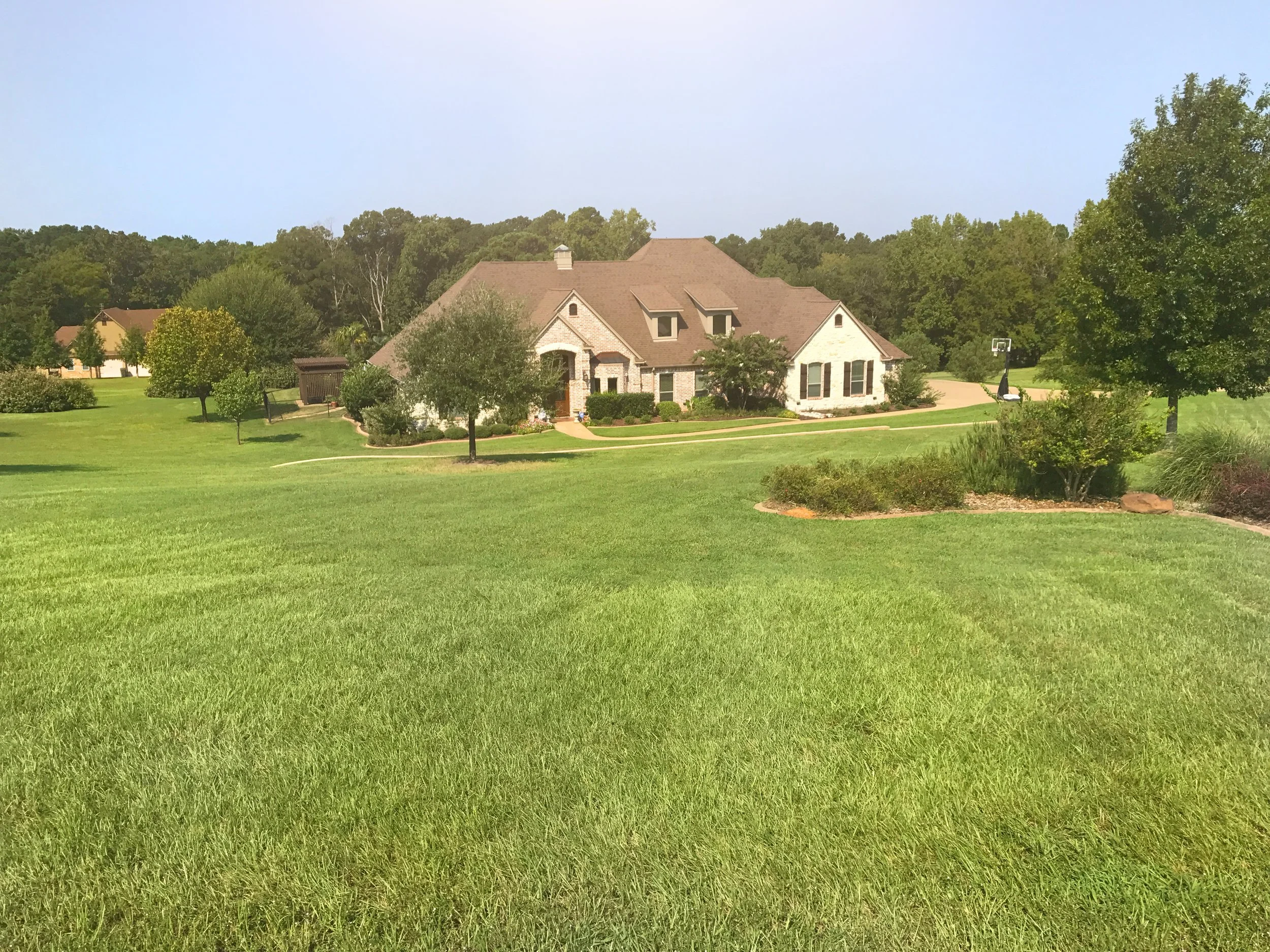 A large house with brick and white siding walls, surrounded by a well-maintained green lawn and trees, on a sunny day.