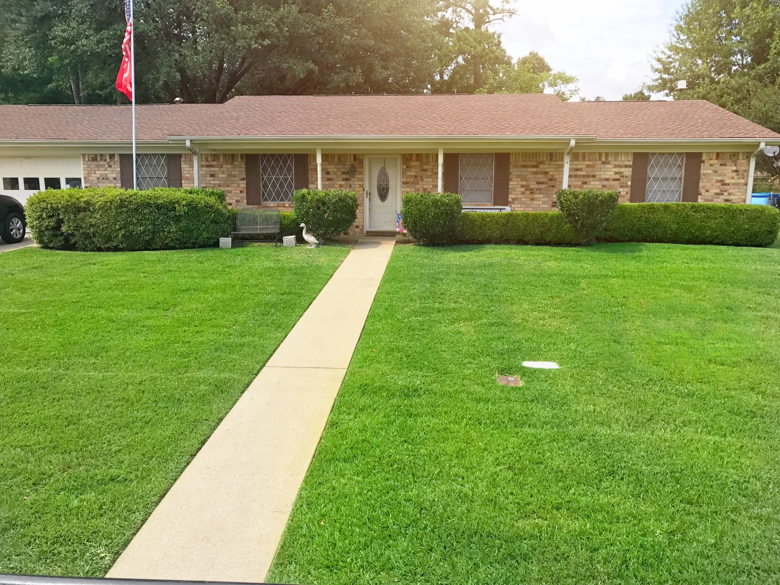 Single-story brick house with a red-tiled roof, green shrubs, and a well-maintained lawn. A straight walkway leads to the front door. There is a white duck on the lawn near the house, and a flagpole with an American flag on the left side of the image