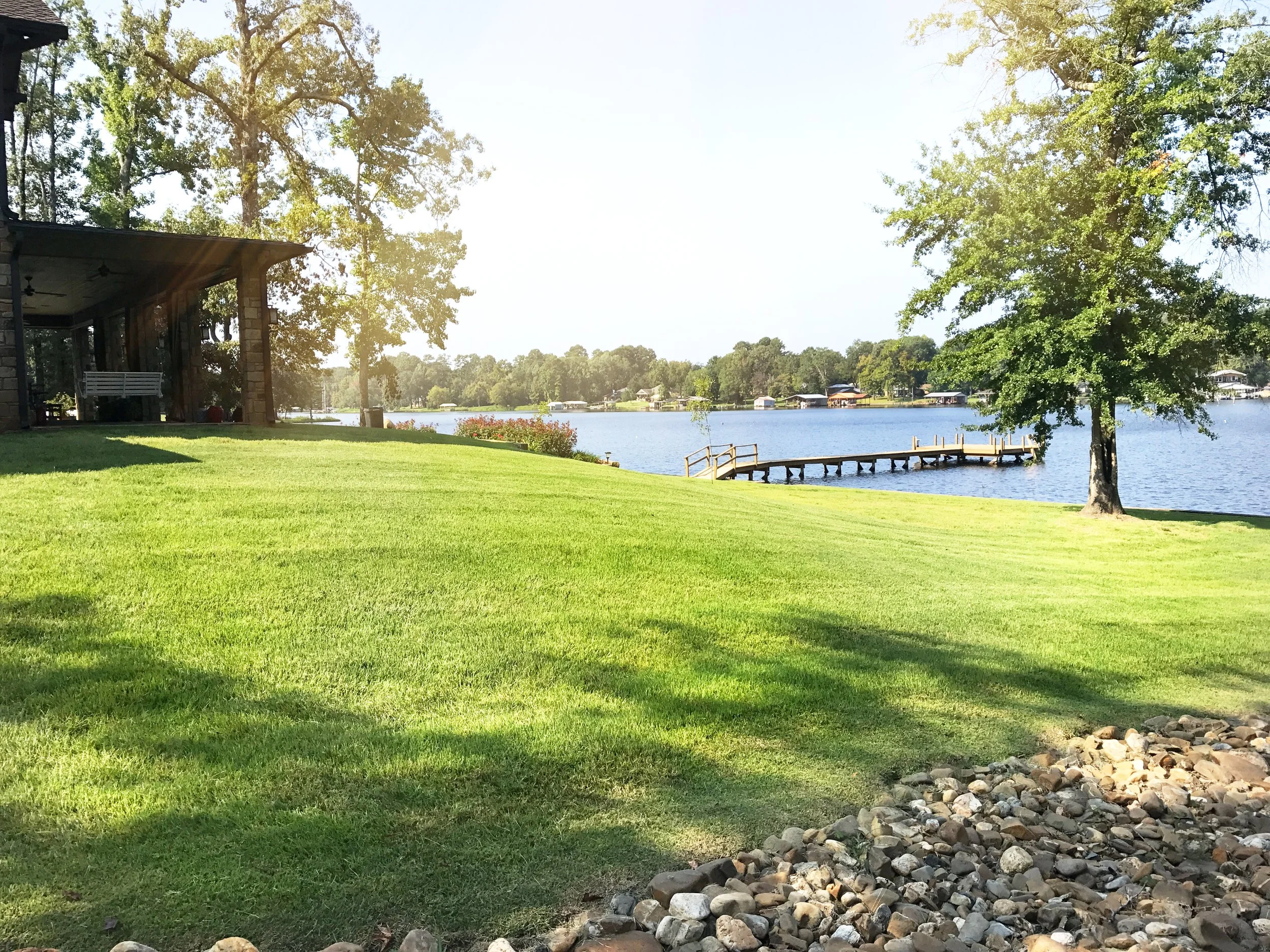 A lakeside scene with a grassy lawn, a dock extending into the water, trees, and a pavilion on the left with a bench underneath, under bright sunlight.