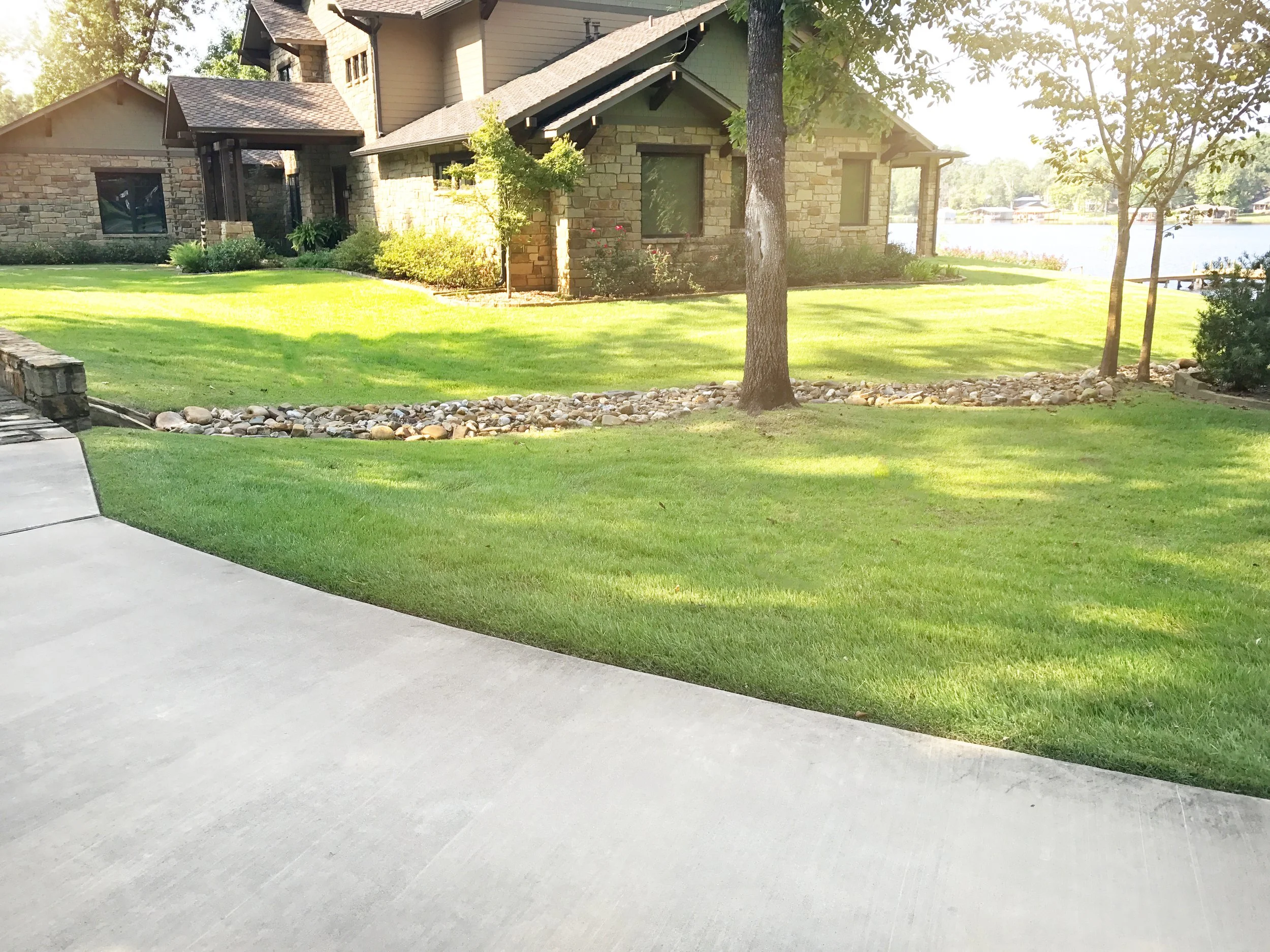 A residential property with a well-maintained lawn, trees, and a stone house near a body of water.