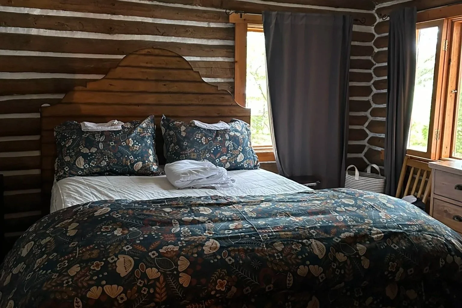 A rustic bedroom with a wooden bed and floral bedding, dark curtains, and windows showing greenery outside.