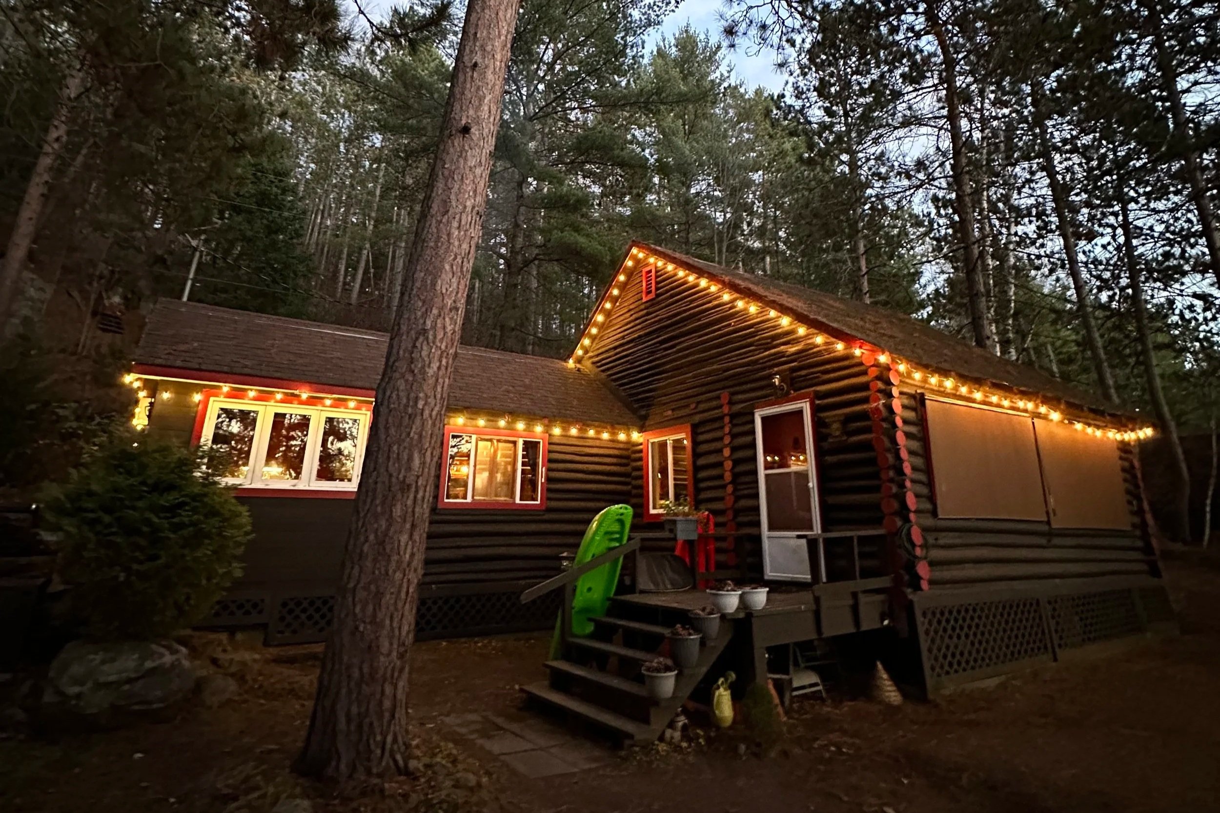 A cozy log cabin decorated with string lights at dusk, surrounded by tall trees in a wooded area.