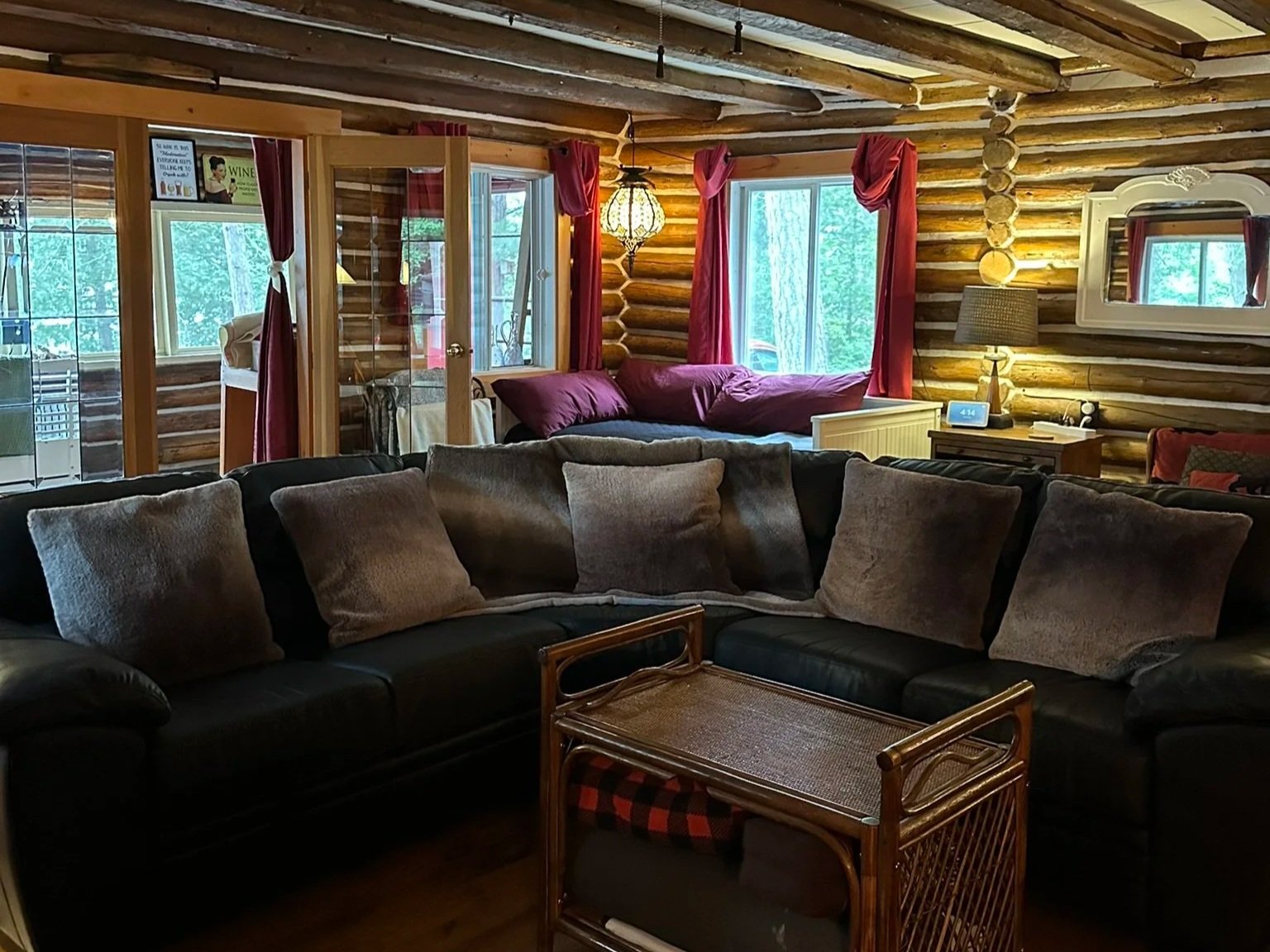 A cozy living room with a large black sectional sofa, beige cushions, wooden floors, and log cabin walls. There is a small wooden side table with a checkered blanket underneath, window with red curtains, and a bed with red bedding in the background. 