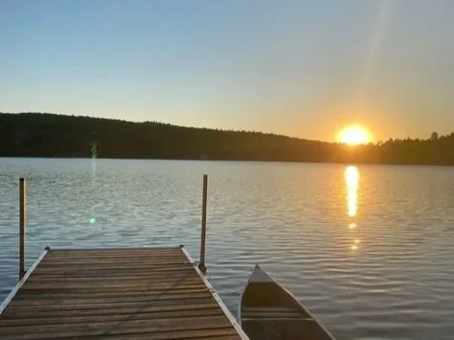 A wooden dock extends into a calm lake at sunset, with a small boat tied to the dock on the right side and a distant tree-lined shoreline in the background.