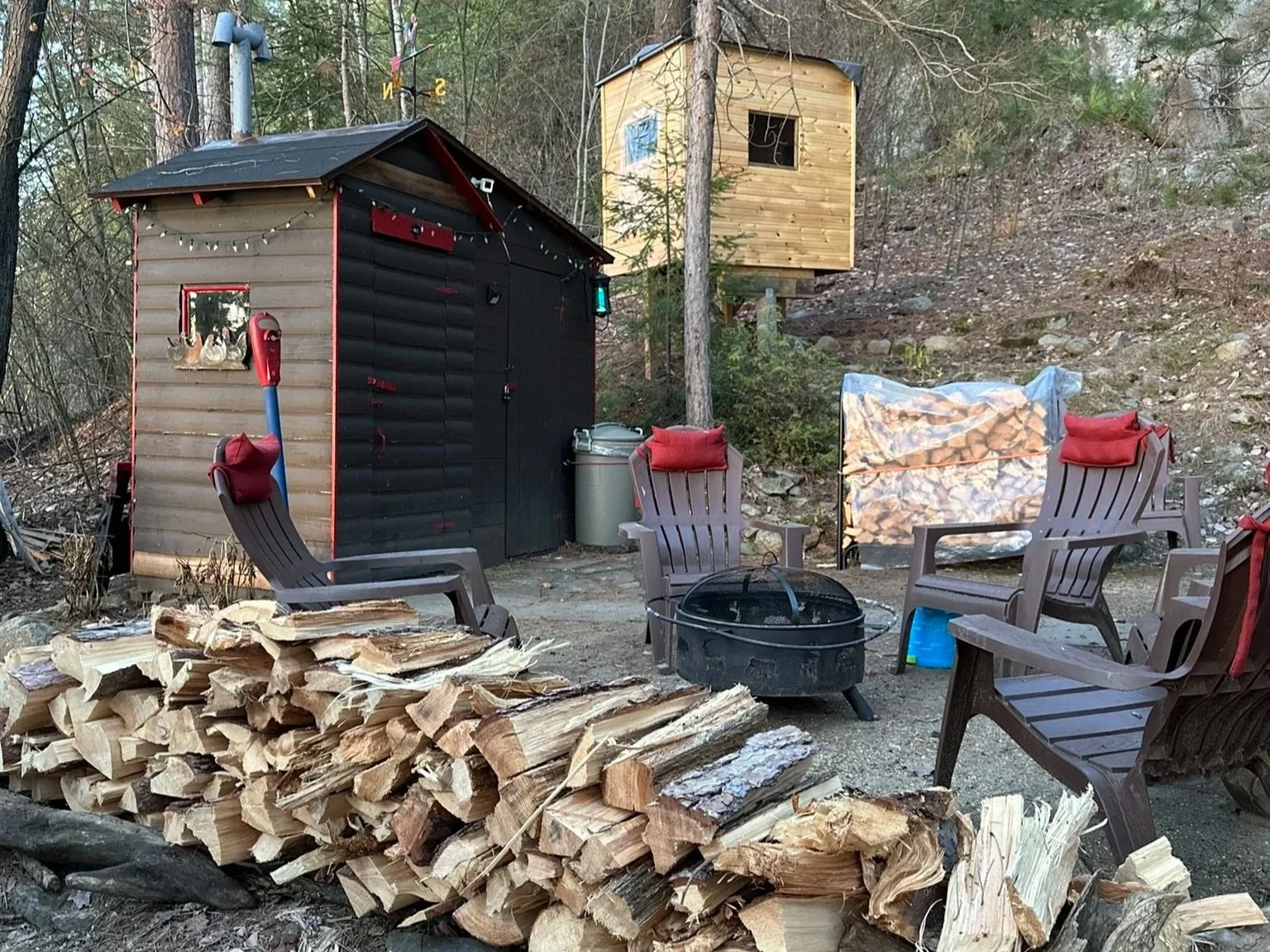 Outdoor scene with a small black shed with red accents and a small window, three chairs, a fire pit, a pile of chopped firewood, and a tiny wooden cabin on stilts among trees in a forested area.