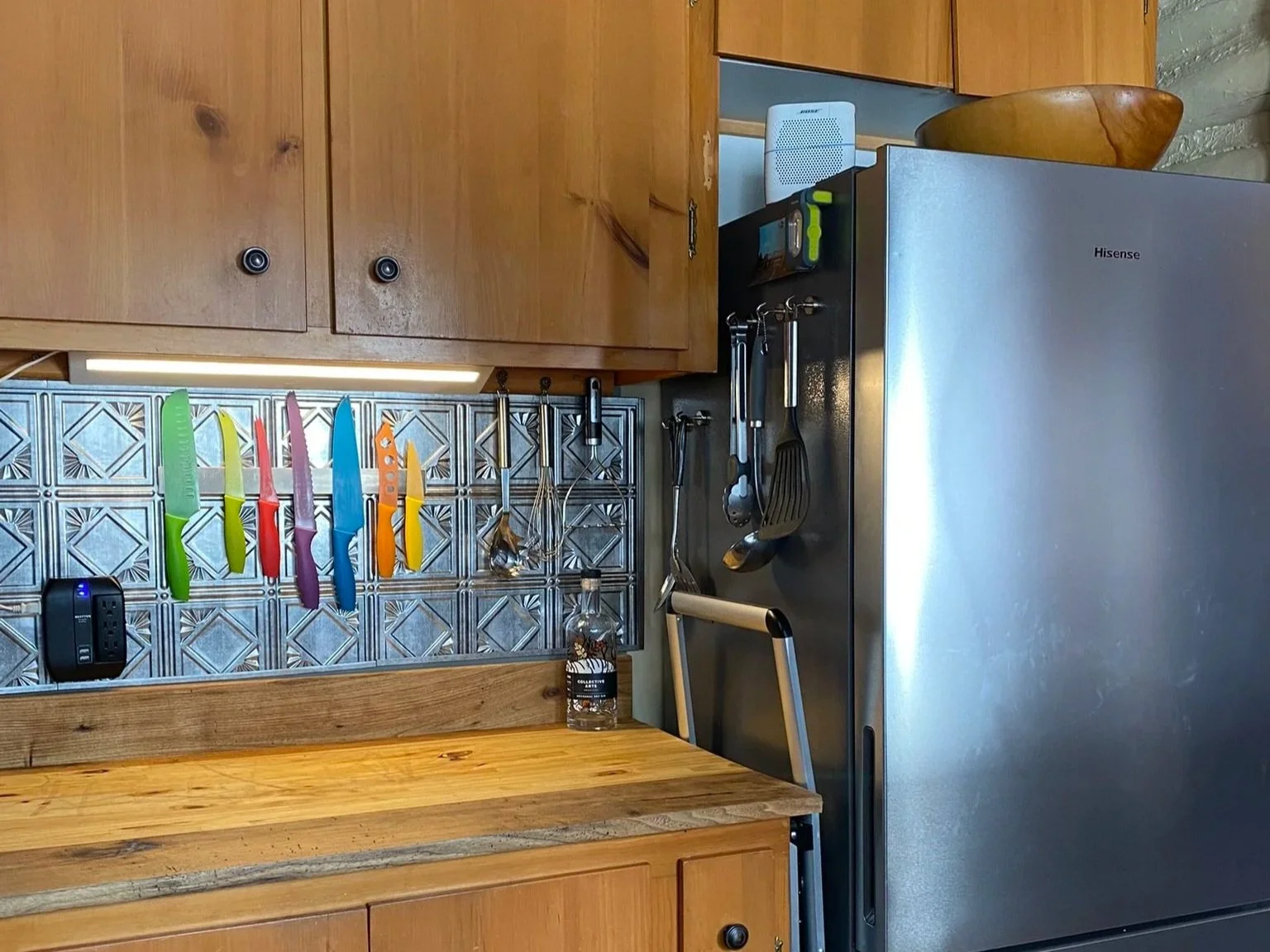 Kitchen corner with wooden cabinets, a stainless steel refrigerator, colorful knives on a magnetic strip, a bottle of cleaning spray, and various kitchen utensils hanging on a wall-mounted rack.