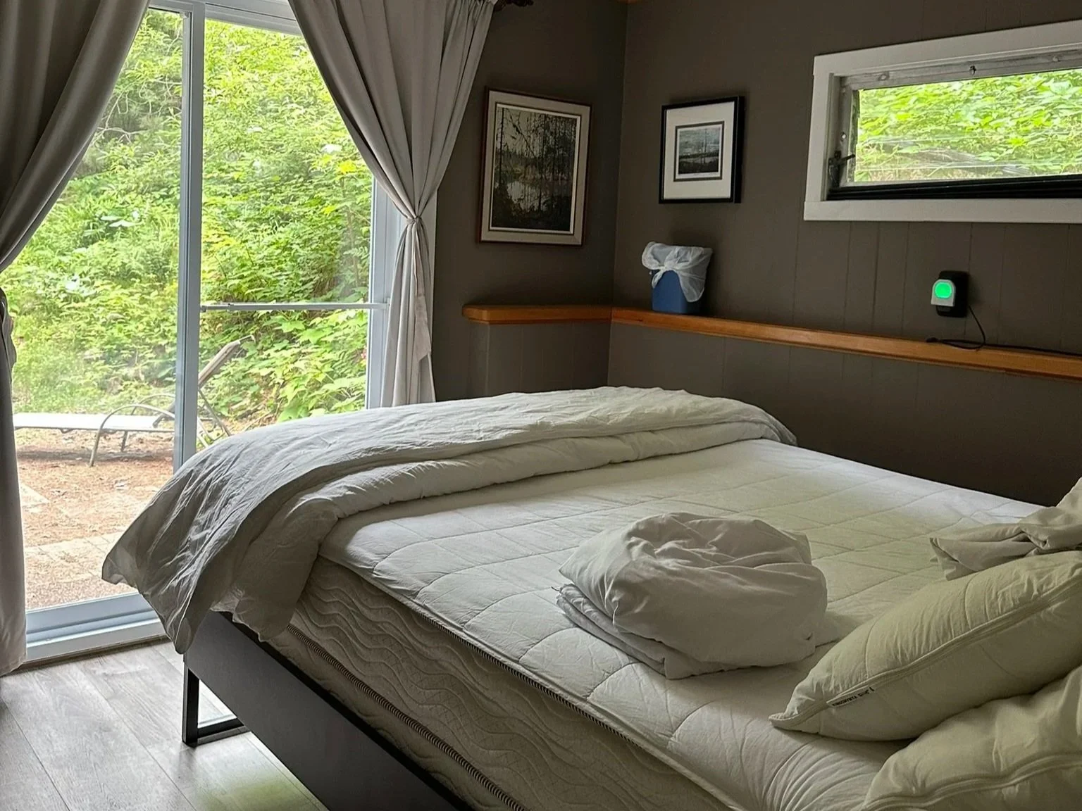 A bedroom with a bed that has rumpled white sheets and pillows, a sliding glass door with curtains leading outside, a window, framed pictures on the wall, a ceiling fan, and a small shelf with a tissue box and a device with a glowing green light.
