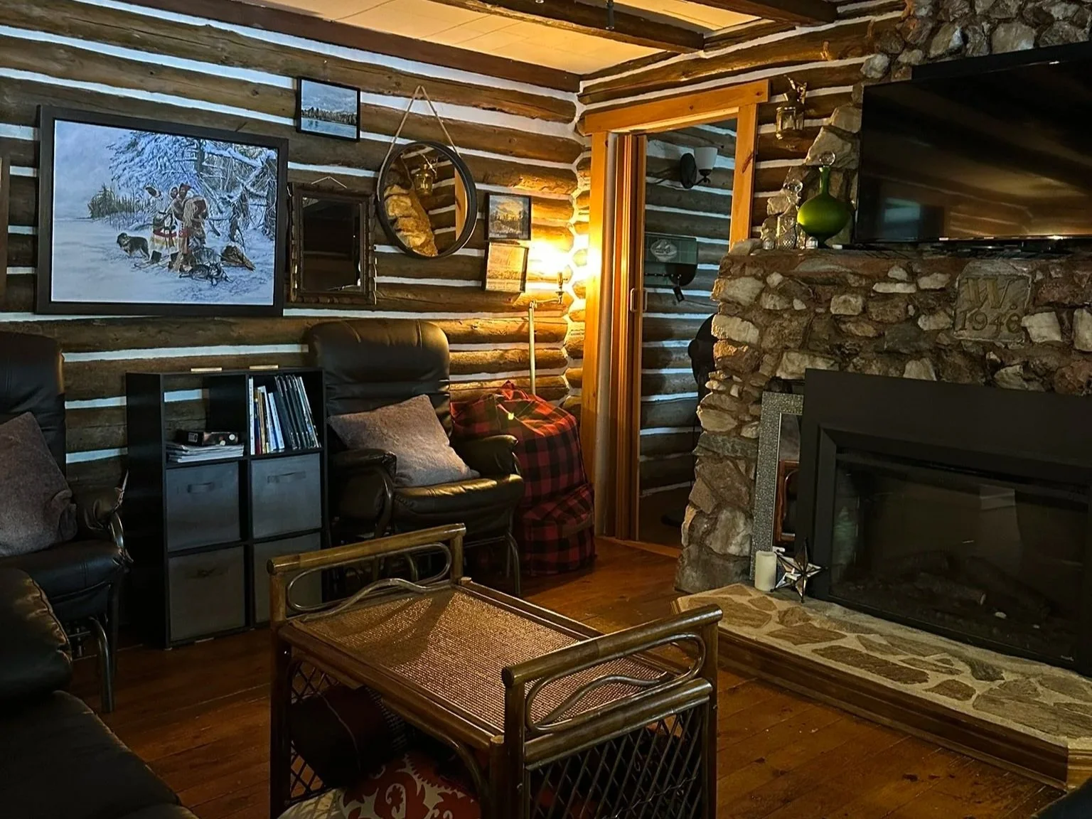 Cozy living room with log cabin style decor, featuring a stone fireplace, wooden beams on the ceiling, leather chairs, a small table, wall art, and a sliding door leading outside.
